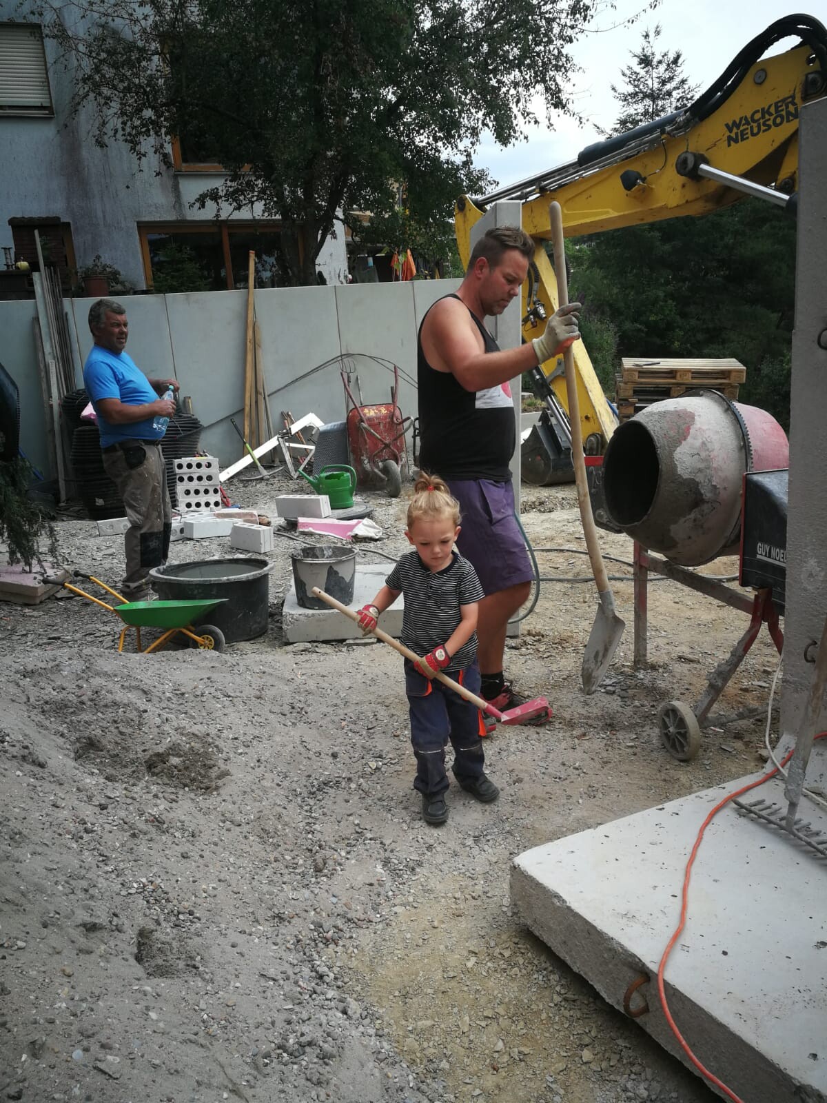 Construction site with man and child; child shoveling soil, concrete mixer in the background.