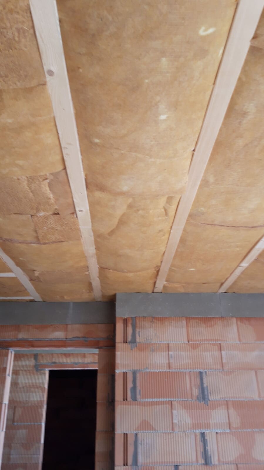 Unfinished interior with ceiling insulation between beams and raw brick wall section