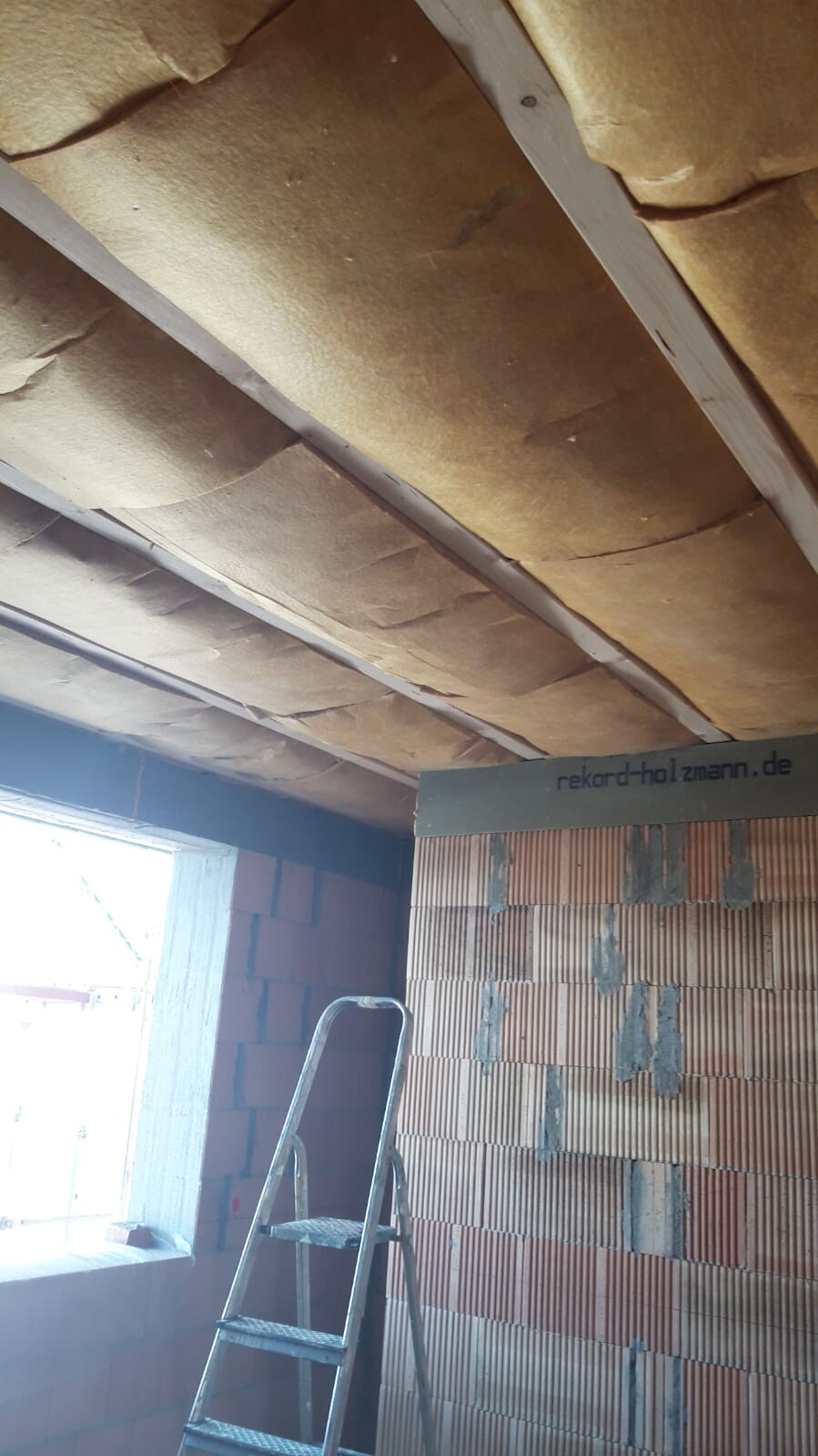 Attic ceiling insulation between beams; ladder and raw brick wall visible