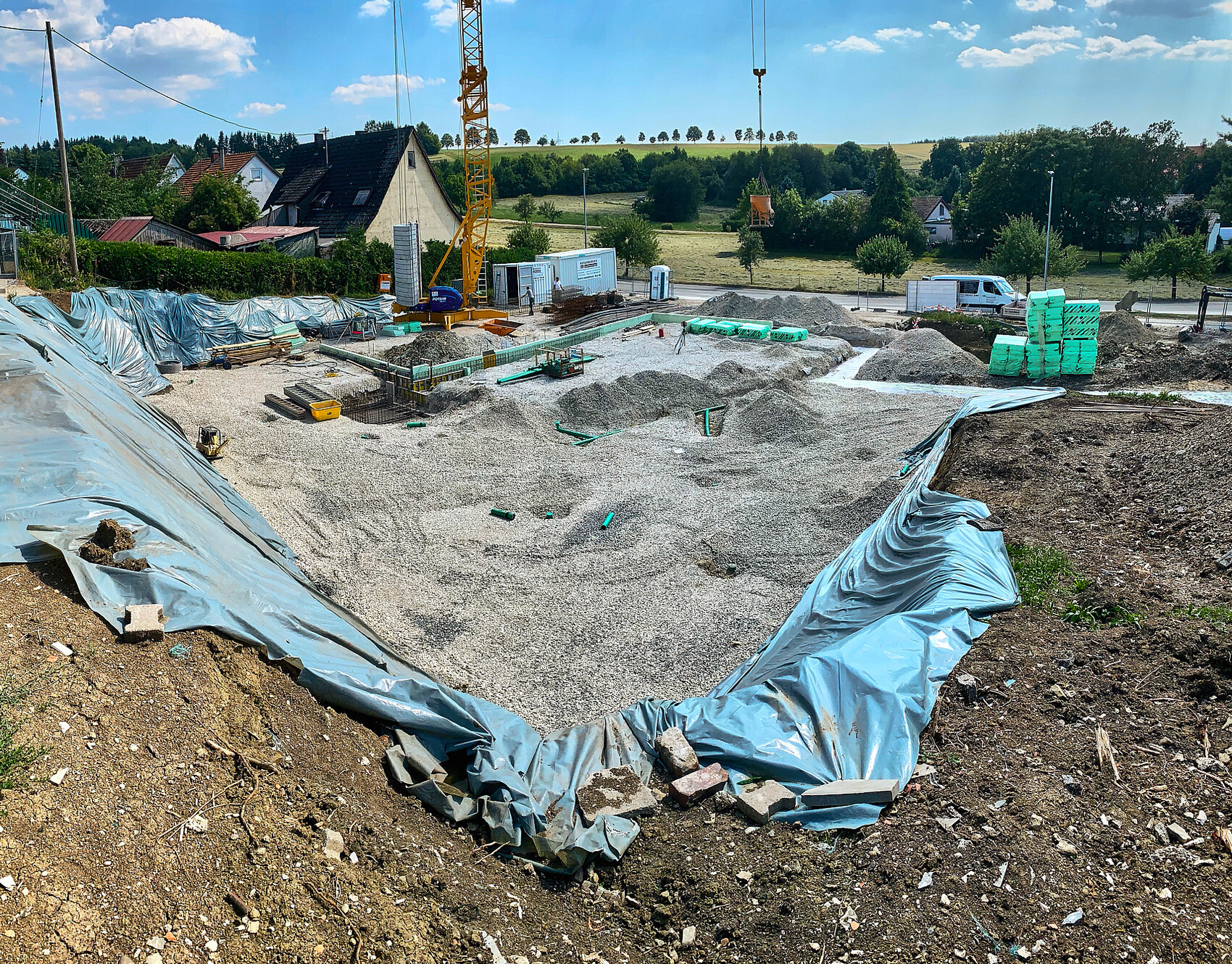 Große Baustelle mit Kran, Kieshaufen, Baugeräten und grünen Materialien, Vorstadthäuser.