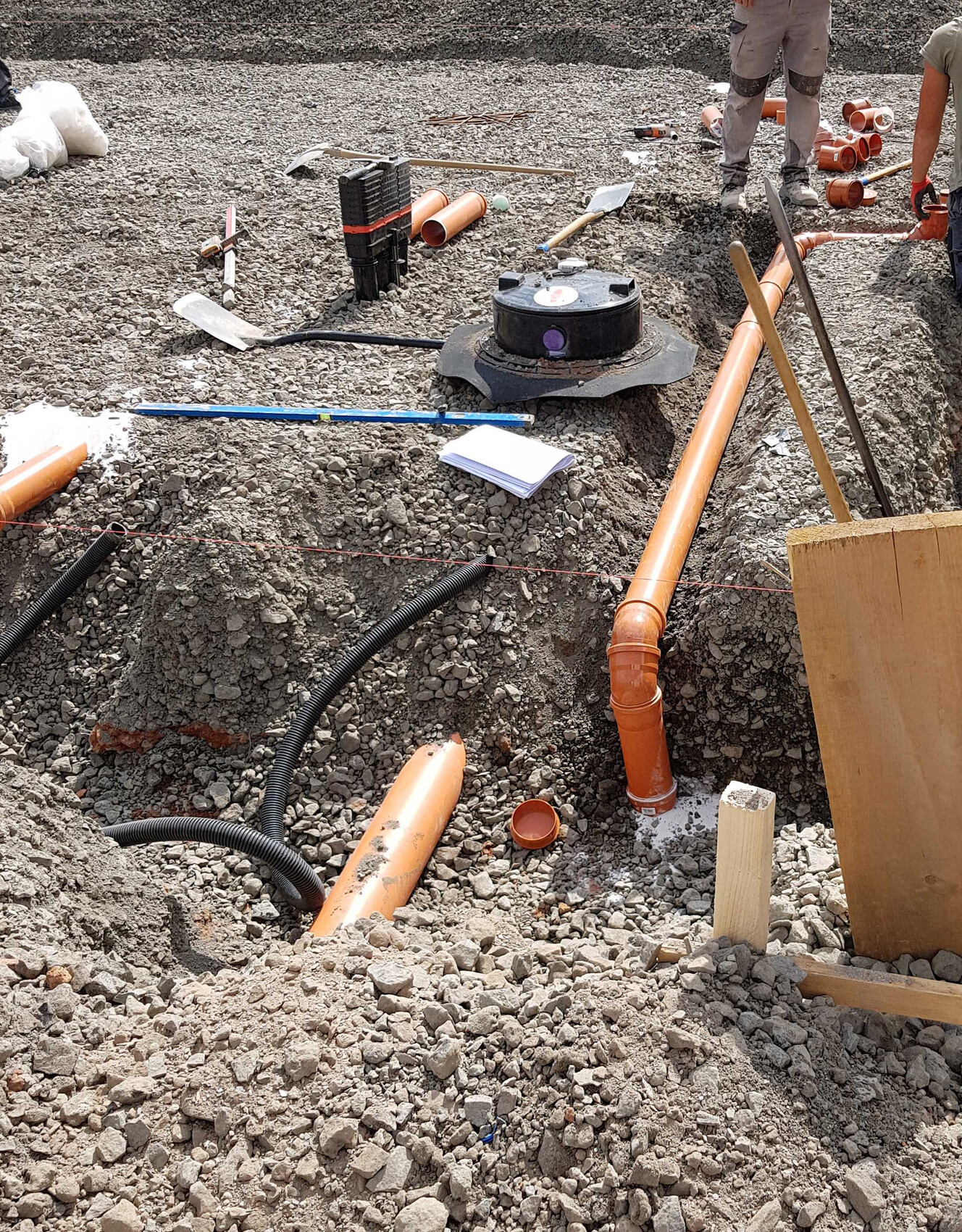 Baustelle mit Grube, orange PVC-Rohre, Werkzeuge, Schaufeln und Bauarbeiter im Hintergrund