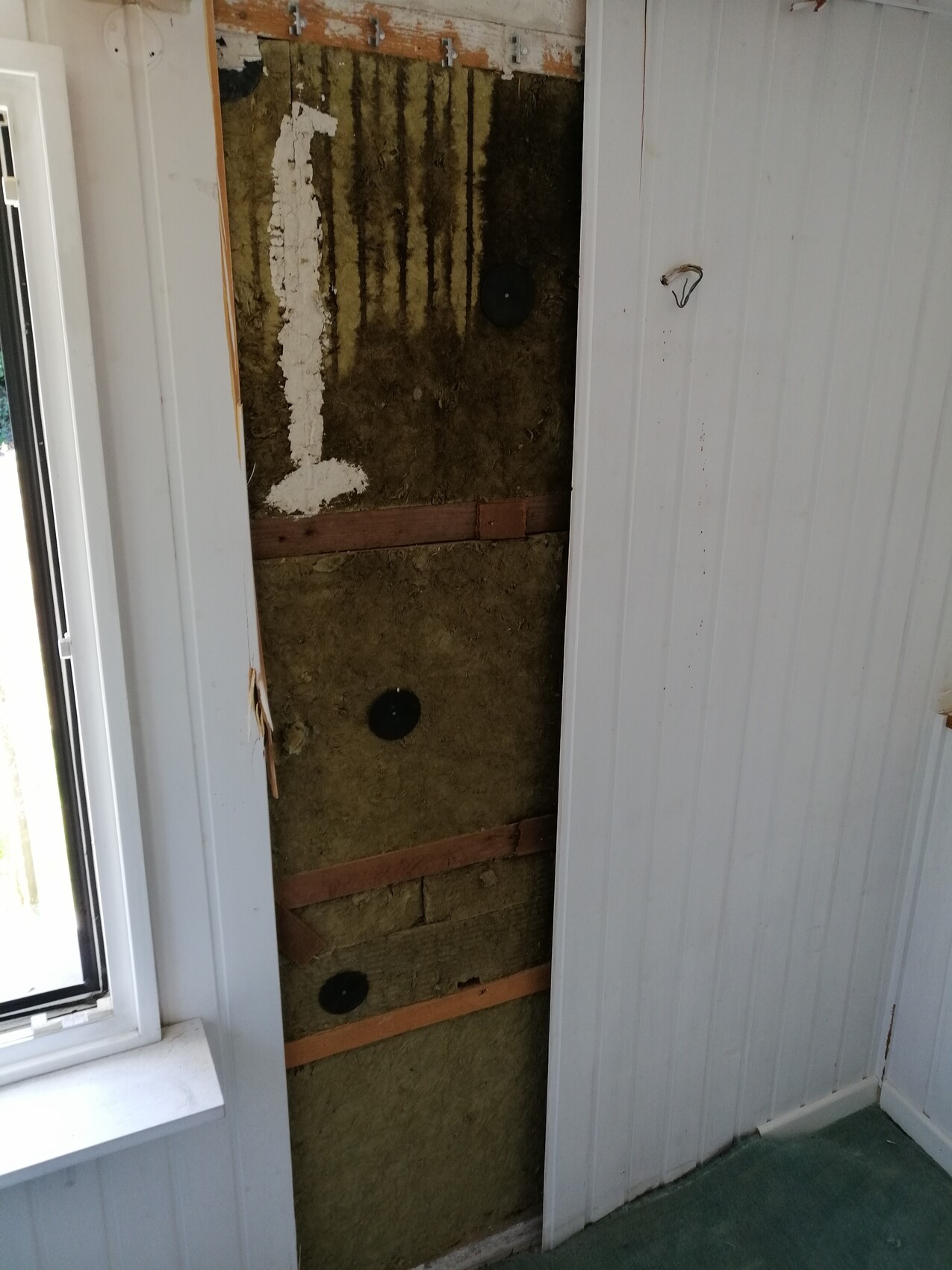 Open interior wall with insulation, wooden battens, plaster remnants, and black round covers.