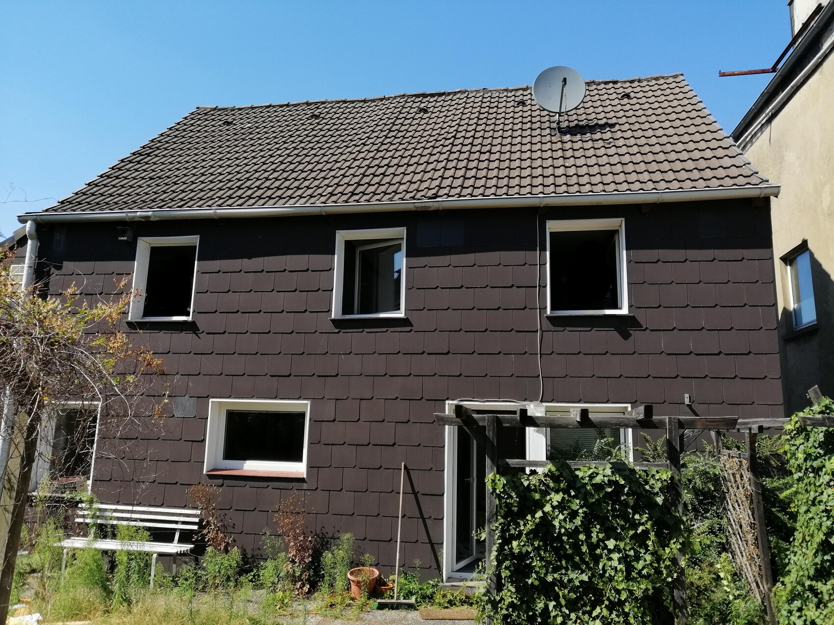 Two-story house with brown shingles, open windows, satellite dish, and lush garden.