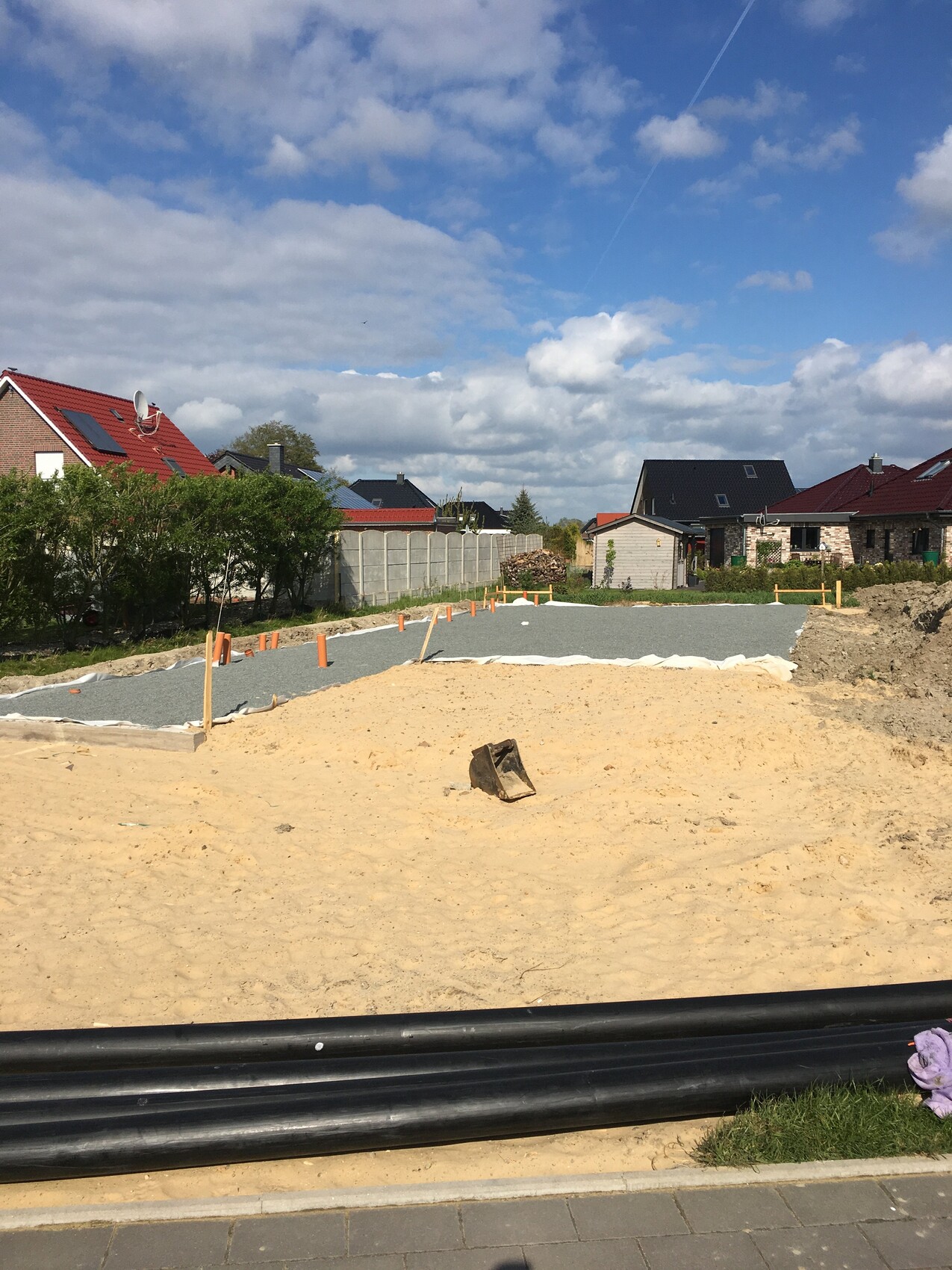 Construction site in residential area: gravel path, sand piles, orange marker poles, houses in the background.