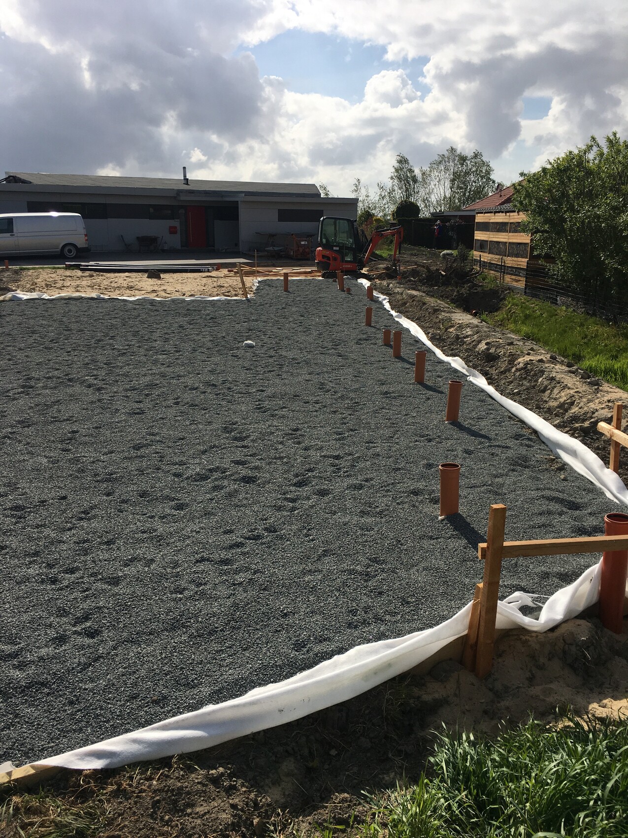 Construction site with gravel base, orange pipes and excavator, geotextile, house in the background.