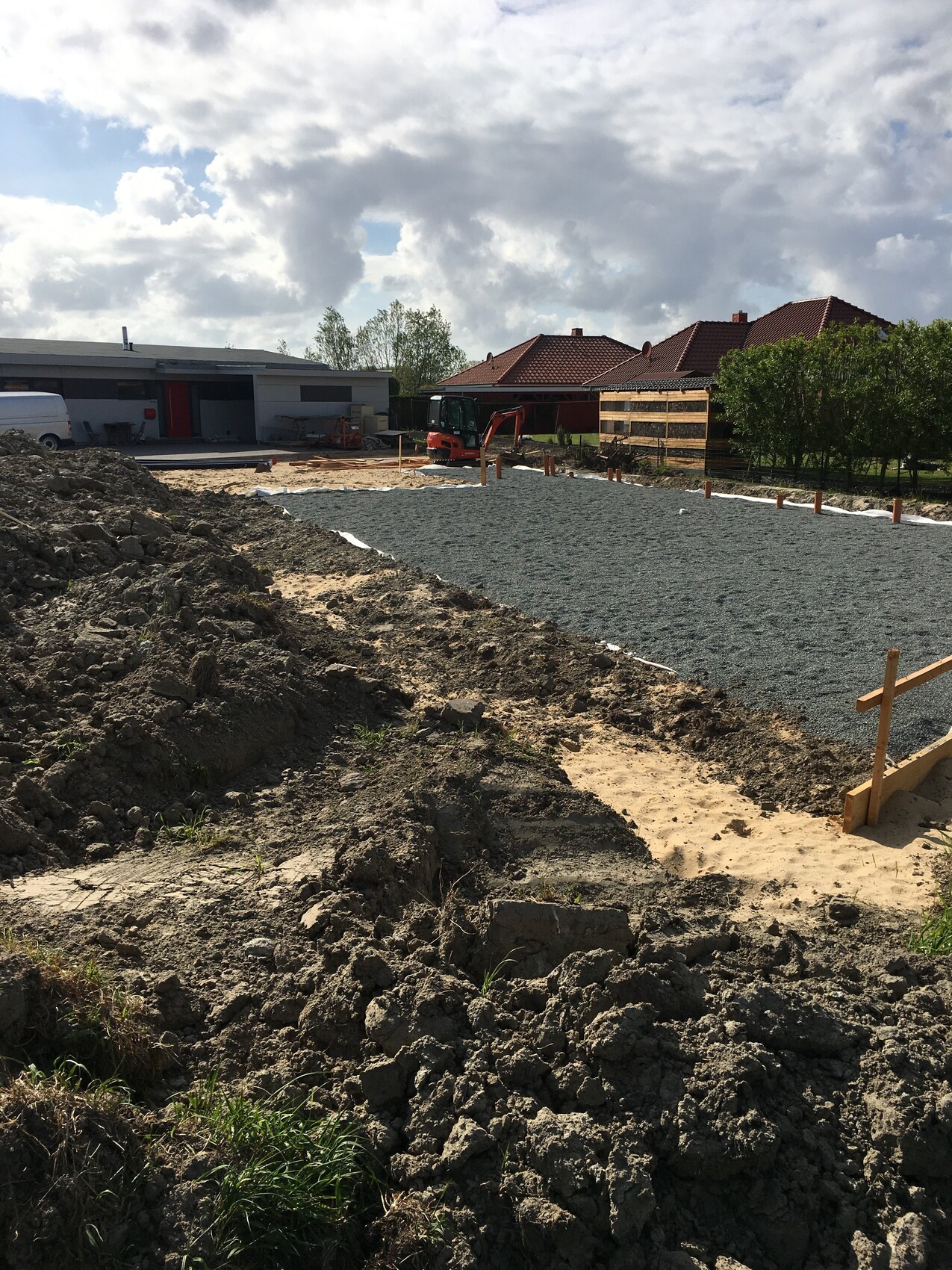 Construction site with gravel surface, earthworks, small excavator and residential houses in the background.