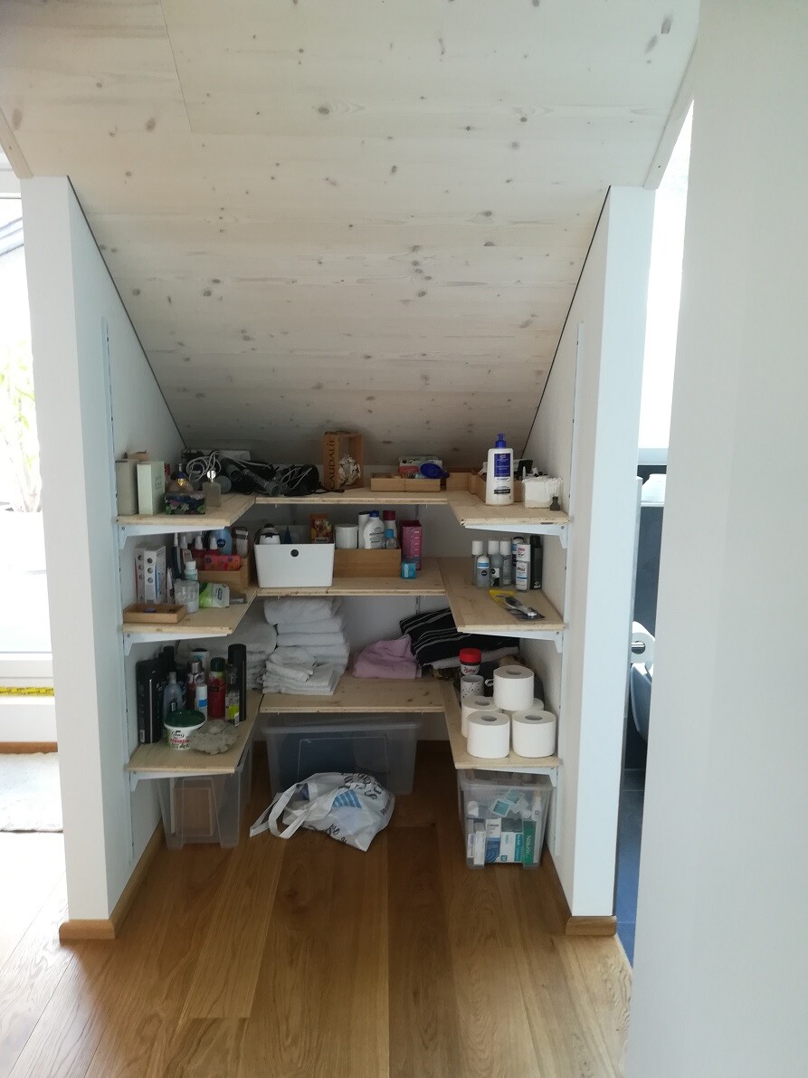 Compact storage space under the sloped ceiling with wooden shelves full of everyday items and toilet paper