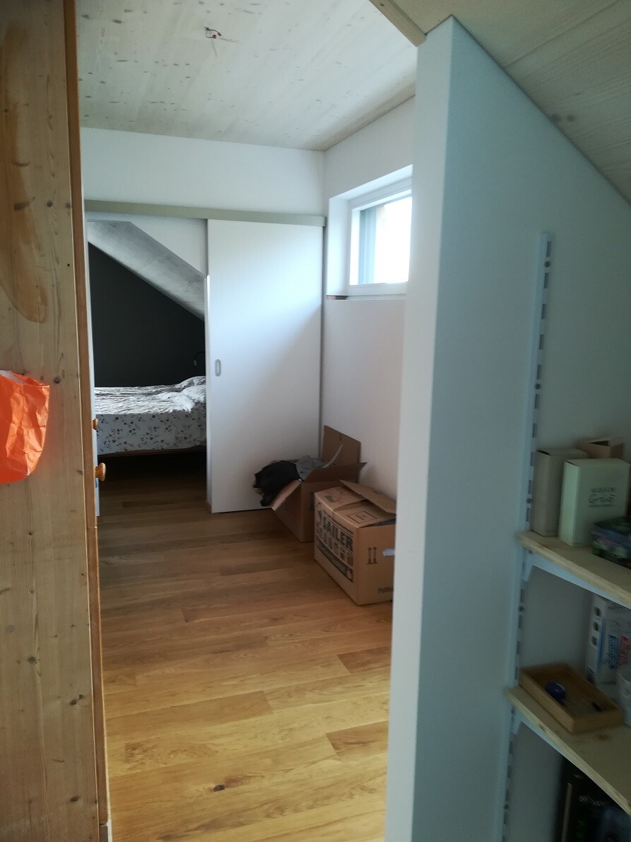Attic room with wooden floor; boxes, orange bag on the left; passage to bedroom with bed.