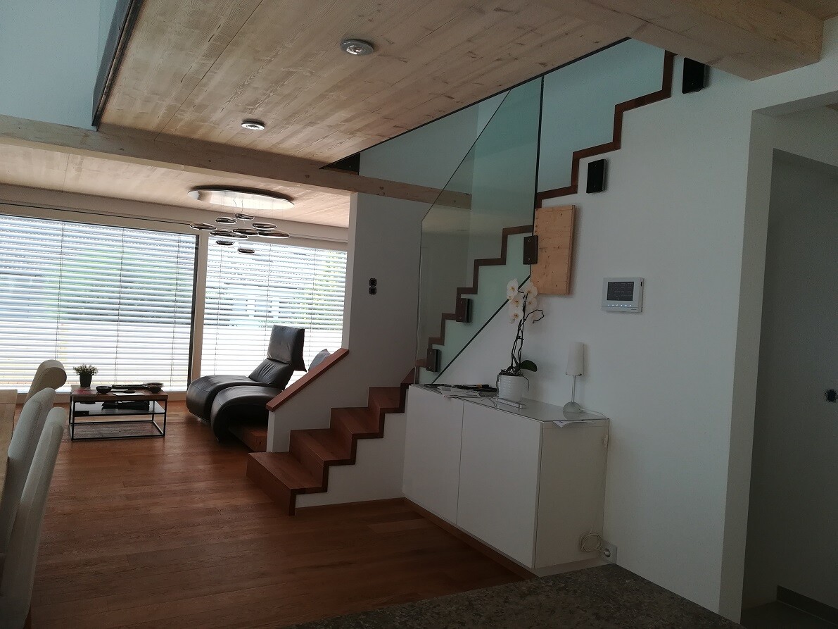 Modern living room with wooden staircase and glass railing, lounge armchair, large windows