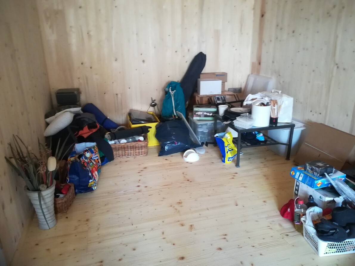 Messy room with wooden walls: boxes, bags, baskets, plants, table full of items.