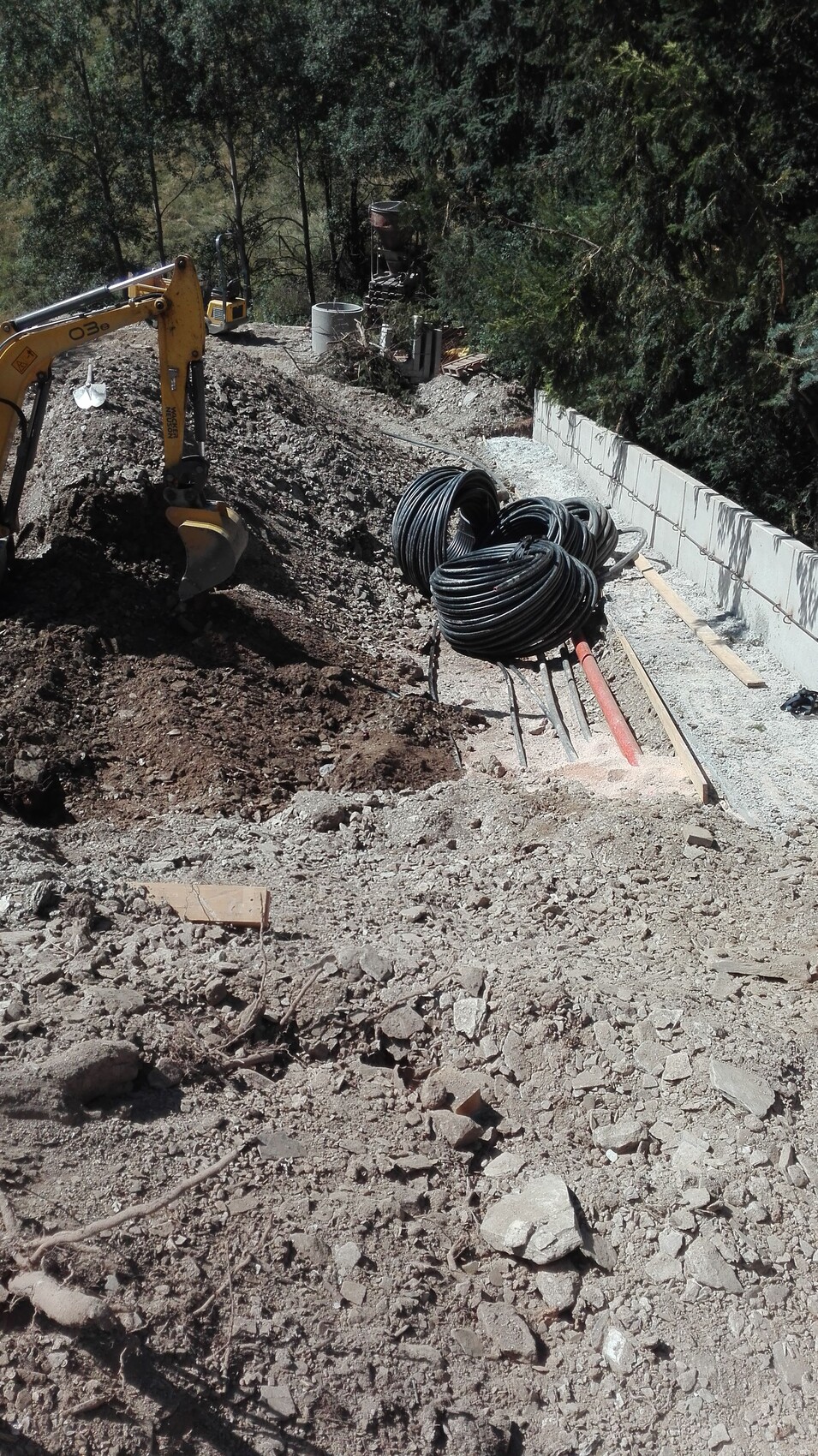 Construction work on a slope: yellow excavator digging earth, several cable drums next to a concrete wall.