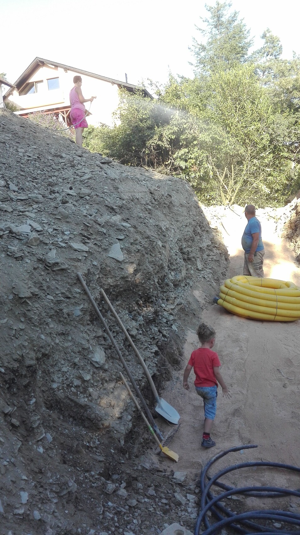 Construction site on a gravel hill: two adults, one child, shovels and yellow hose ready.