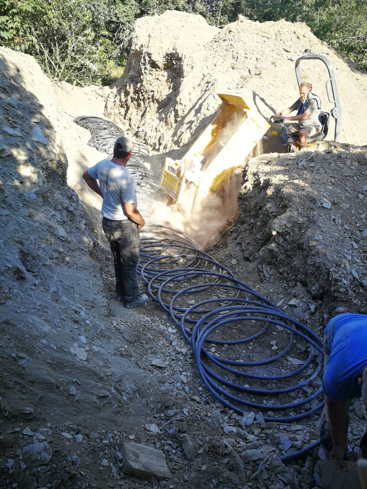 Two construction workers in a gravel pit; yellow front loader unloading, blue hoses lying on the ground.