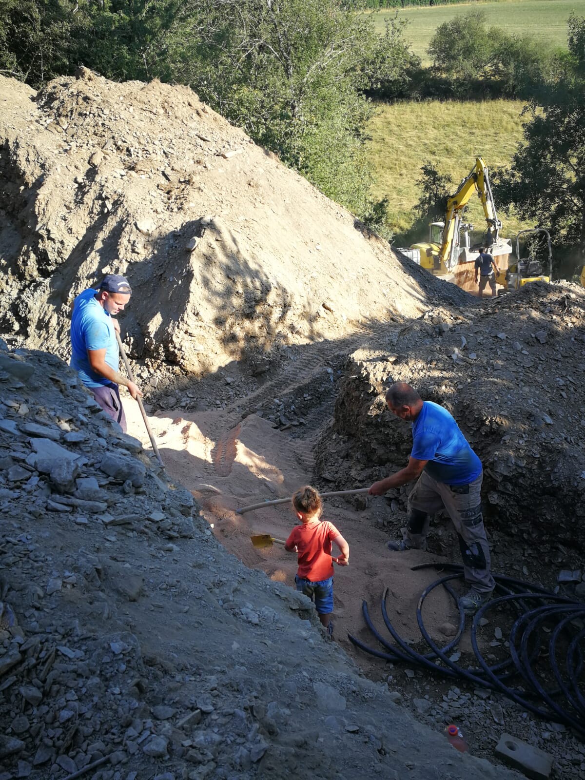 Construction workers shoveling sand into a construction pit; child in red T-shirt watching; excavator in the background.