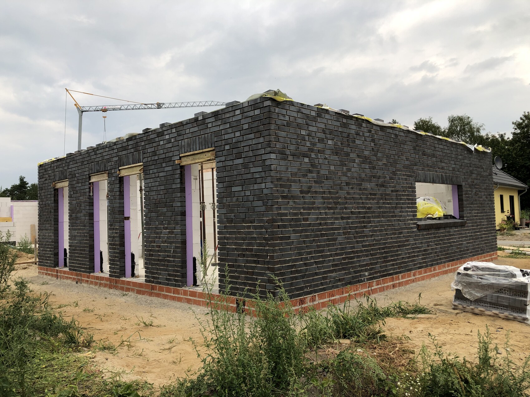 Rohbau eines dunklen Ziegelhauses mit Fenster- und Türöffnungen; Baukran im Hintergrund.