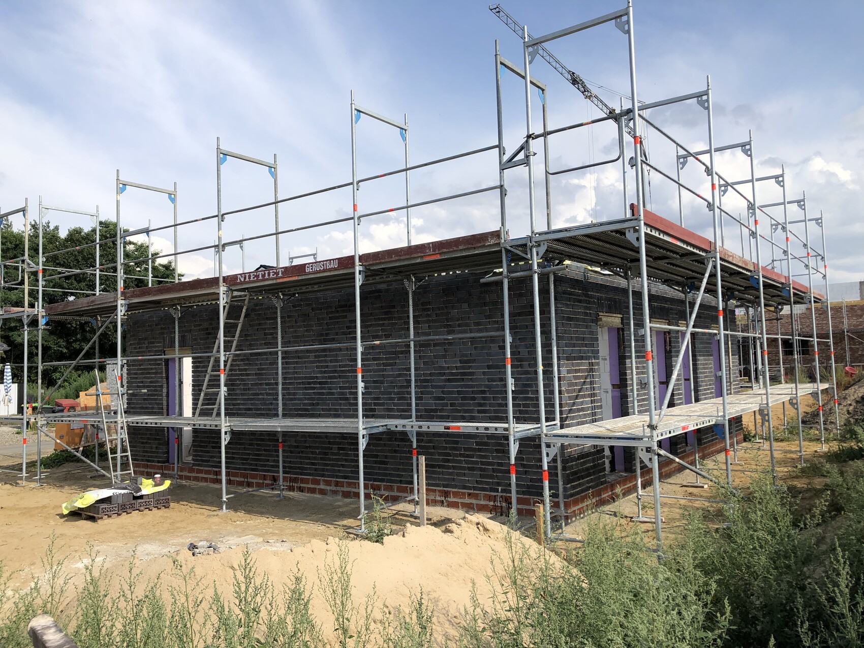 Baustelle mit Gerüst ums Backstein-Gebäude; Bauarbeiten, Sandboden und Pflanzen im Umfeld.