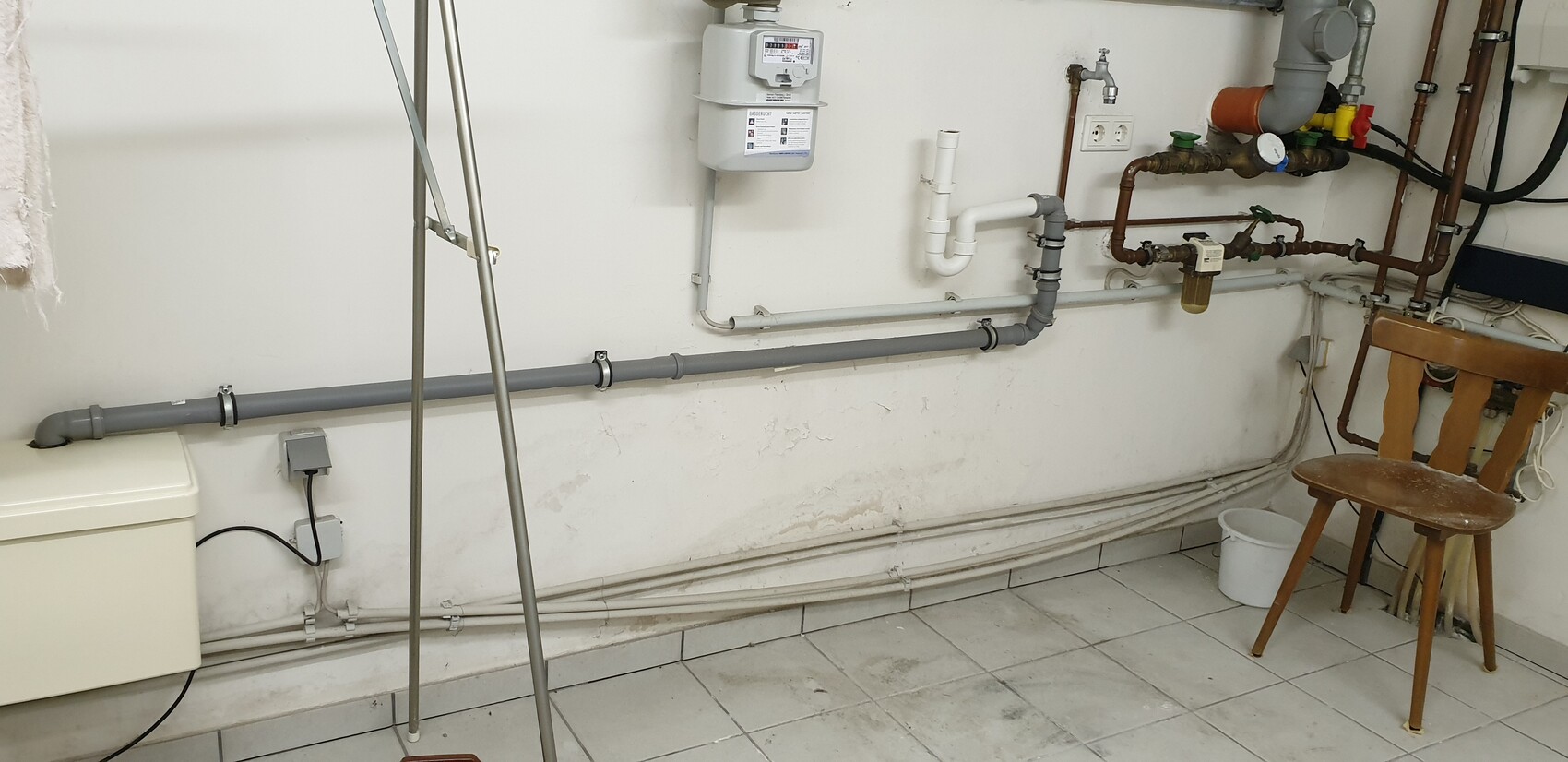 Basement room with gas meter, pipes, valves, chair, and bucket.