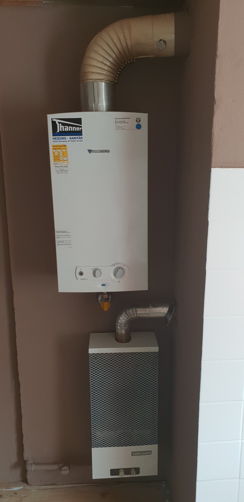 Gas boiler on the wall with curved flue pipe; below it, heating device with metal grille front.