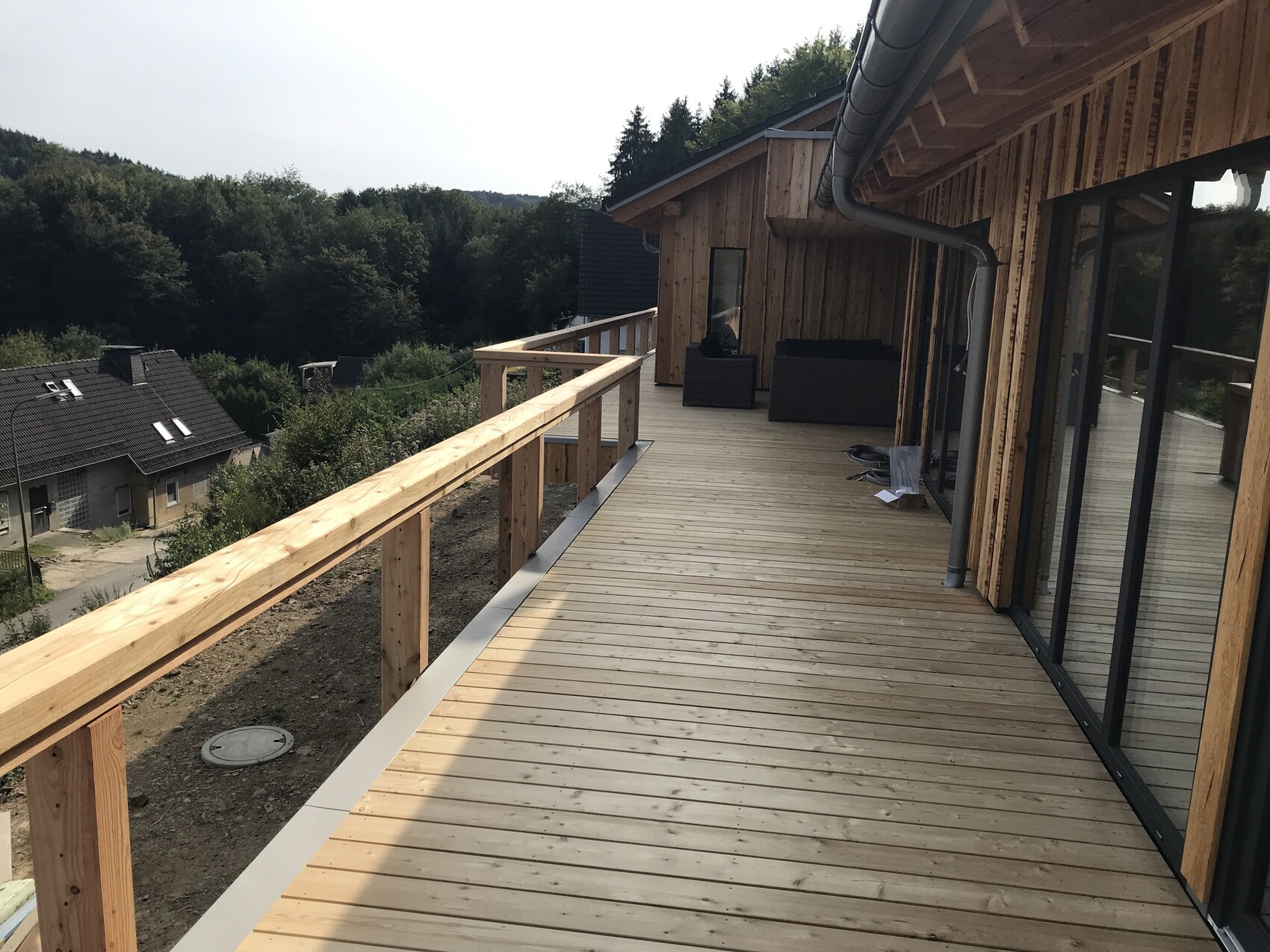 Wooden terrace at the house with railing, glass doors, outdoor furniture, forest view.