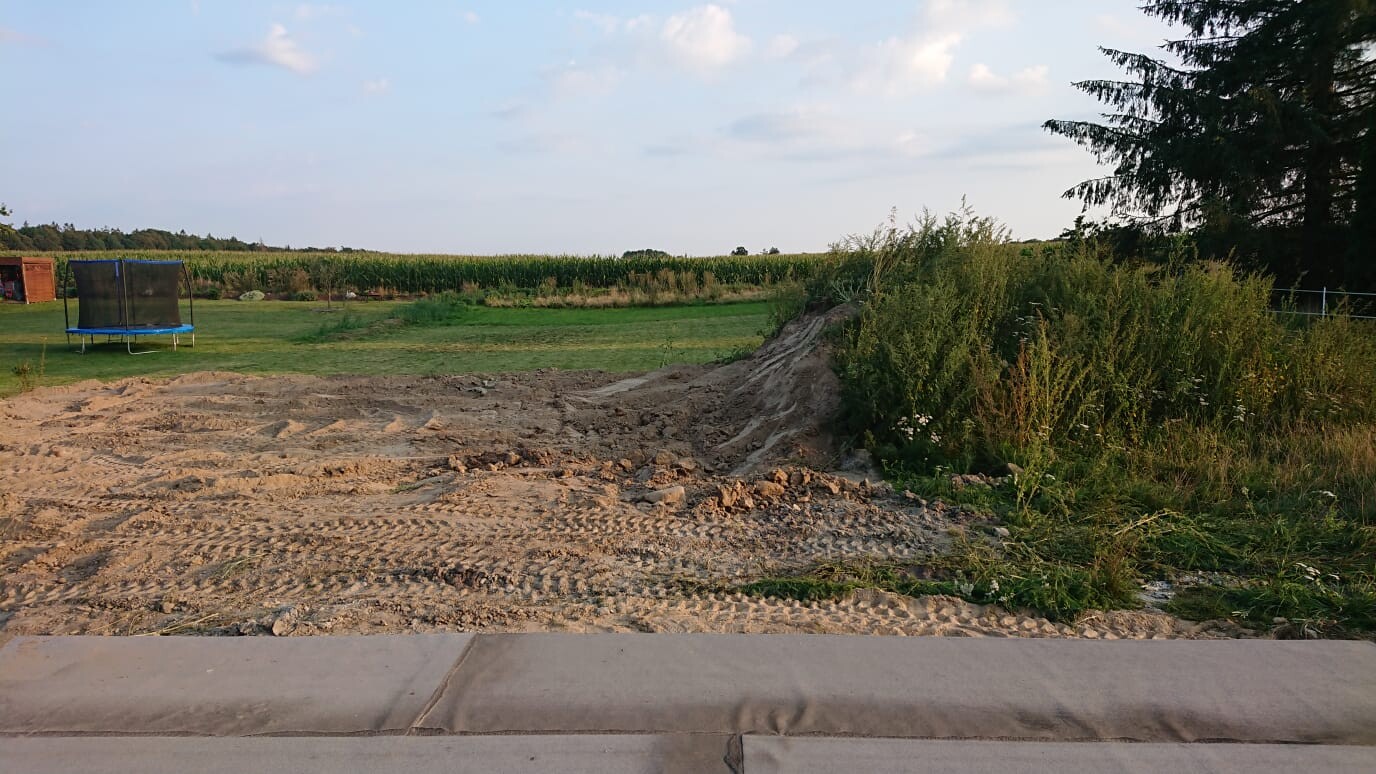 Construction site outdoors with earthworks, unpaved ground, grassy area and trampoline in the background
