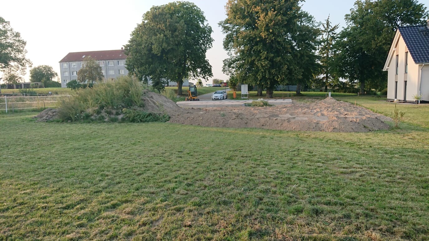 Green field with excavation pit, soil piles, excavator, cars and houses in the background
