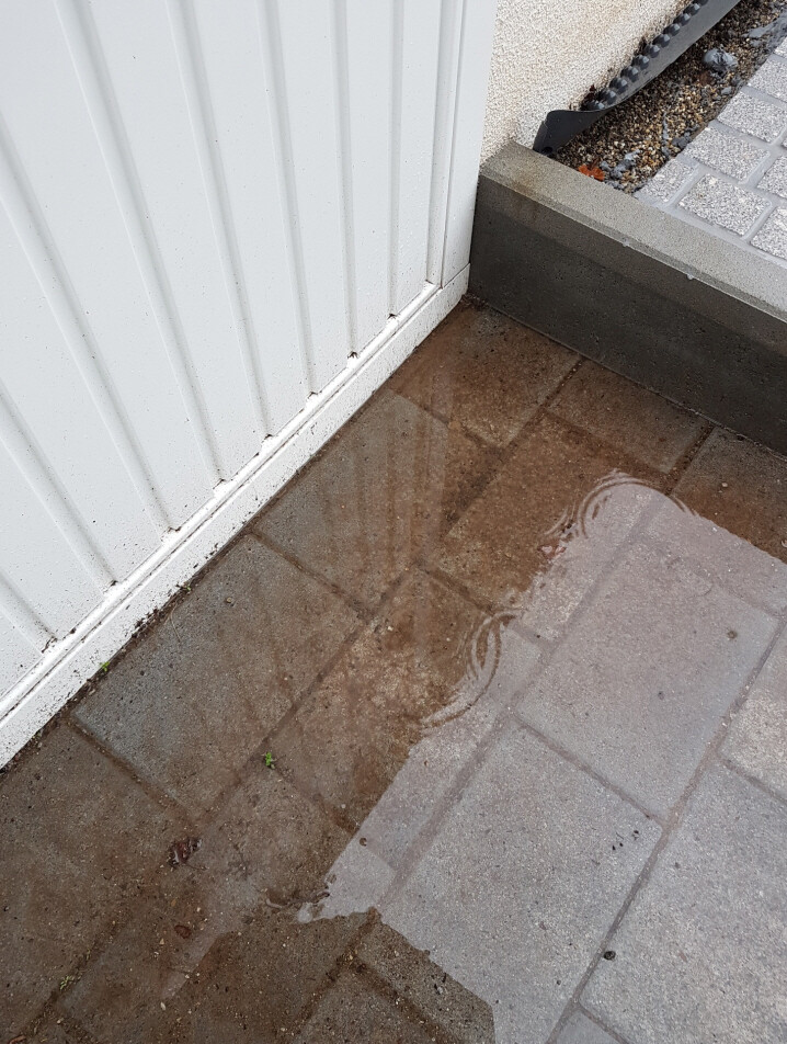 Wasserpfütze auf gepflastertem Boden neben einer weißen vertikalen Außenwand aus Metall.