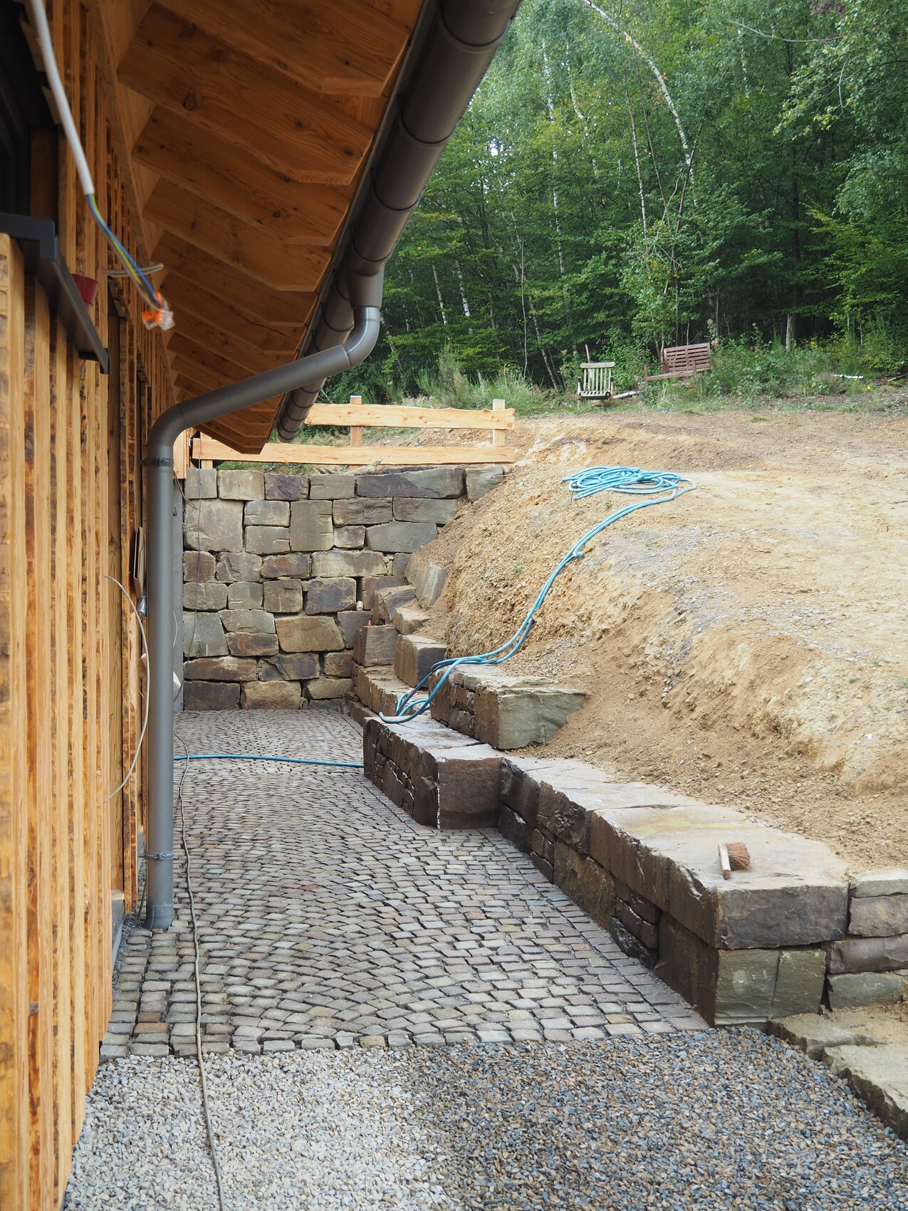 Exterior view of a building site with stone-paved terrace, downpipe, and forest in the background