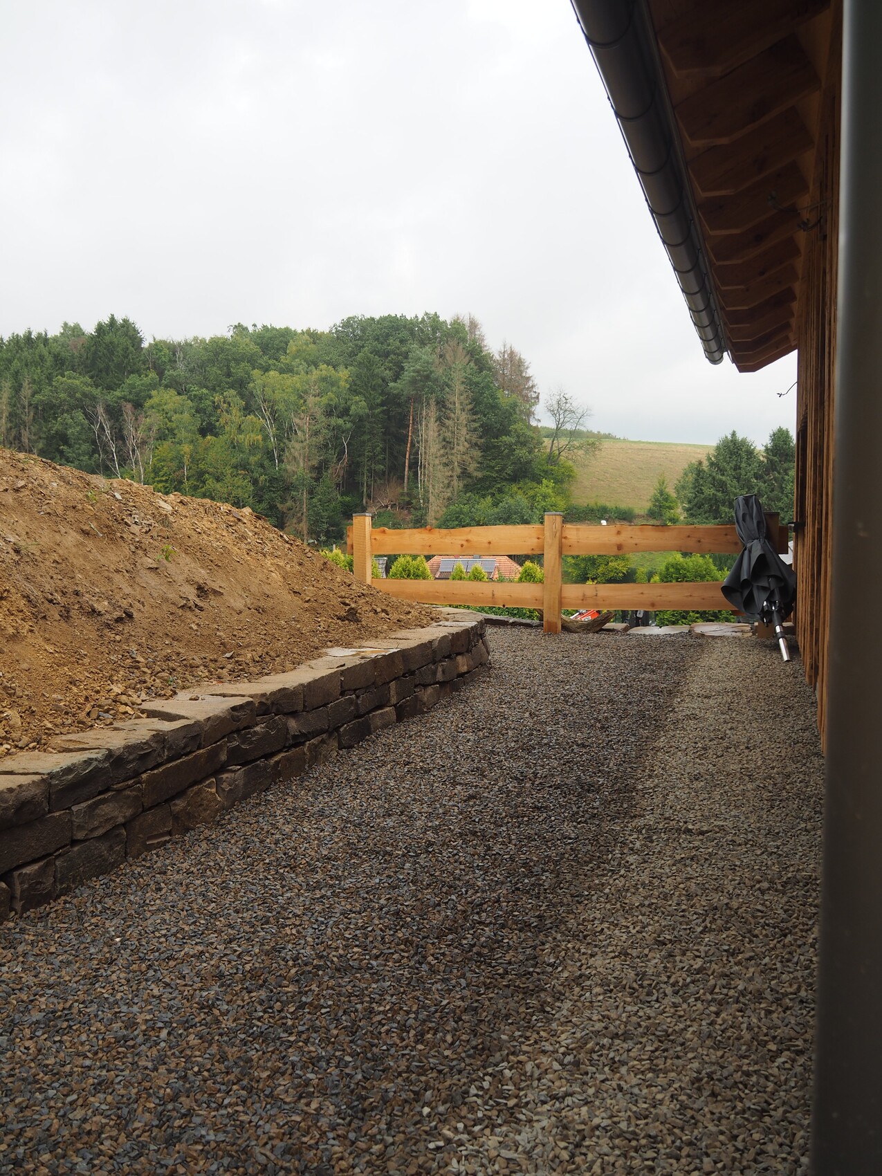 Gravel path next to stone wall, left excavation soil, right house with gutter, wooden fence; forest in the background.