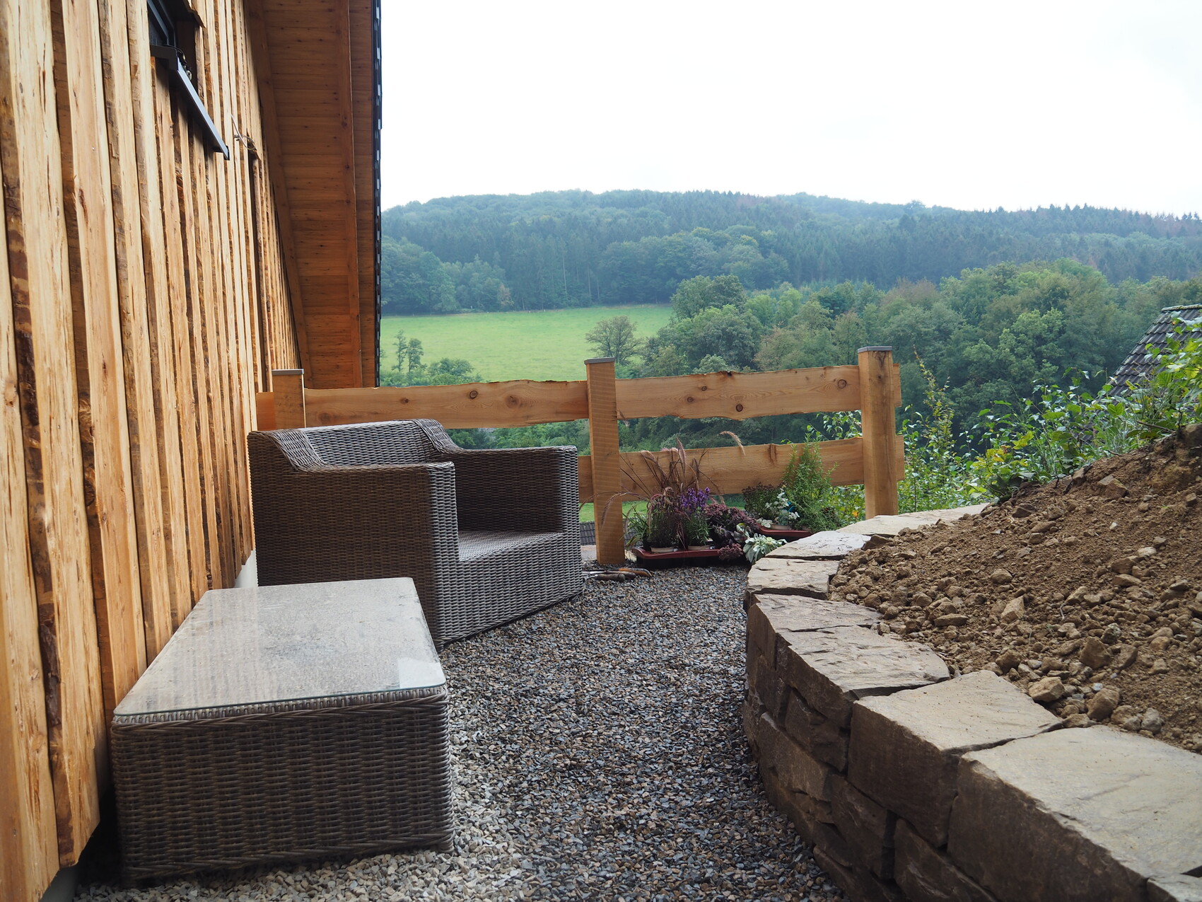 Wooden veranda with rattan chair, stone table, planter boxes; view of fields and forest.