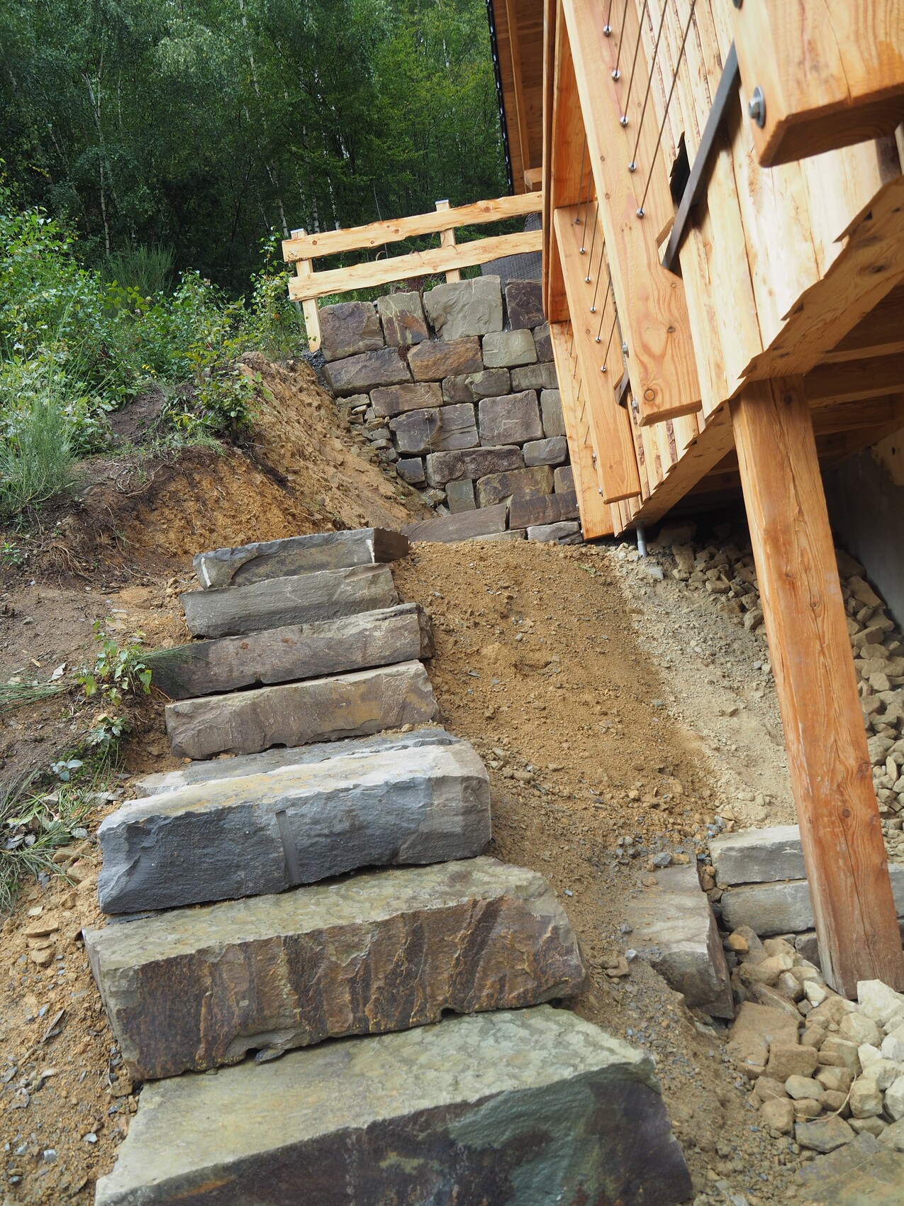 Slope stabilization made of stone with stairs of large blocks next to wooden house, stone wall in the background