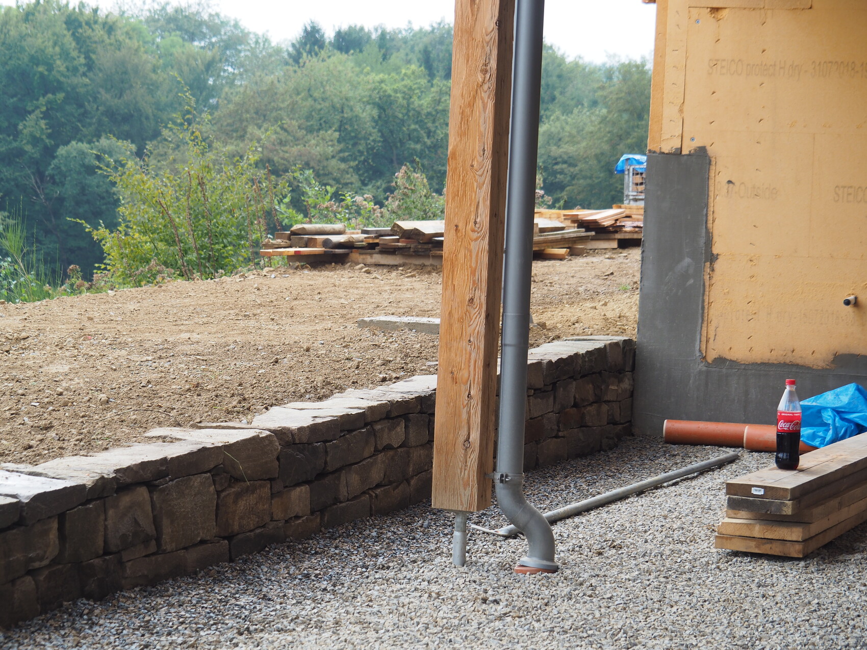 Construction site with stone wall, wooden posts, downpipe, wood stacks and soda bottle.