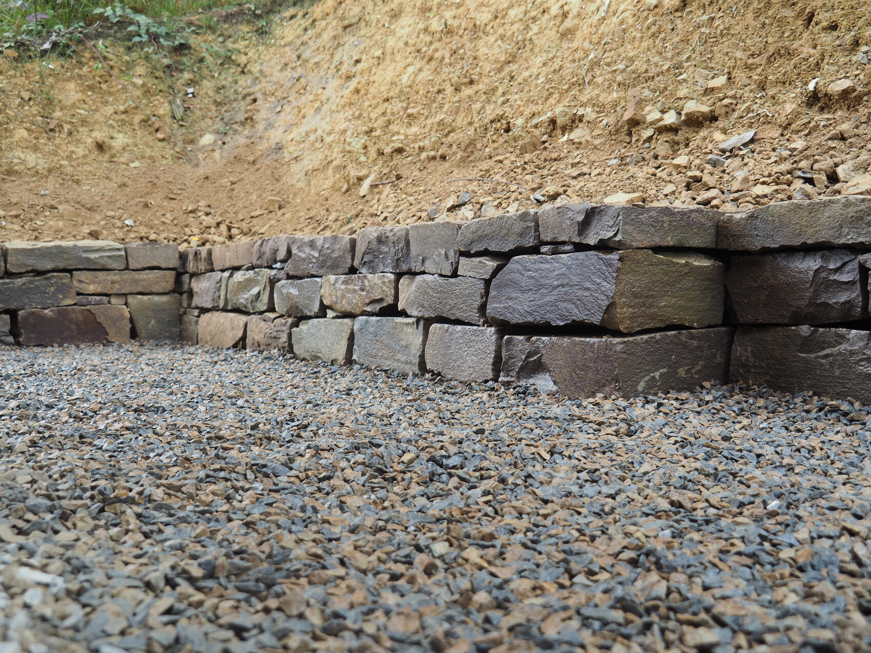 Retaining wall made of coarse natural stones in front of gravel ground and earth embankment.
