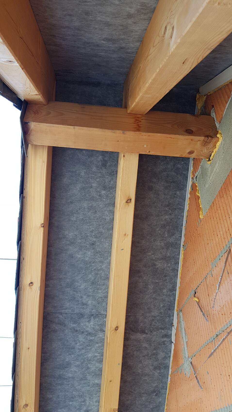 Holzständerwerk mit grauer Dämmung, rechts eine Ziegelwand mit Dichtstoff.
