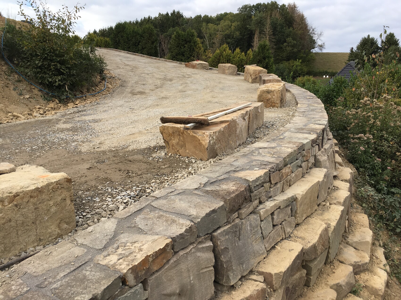 Natural stone wall on a slope, unpaved driveway, gravel and stone block with tools.