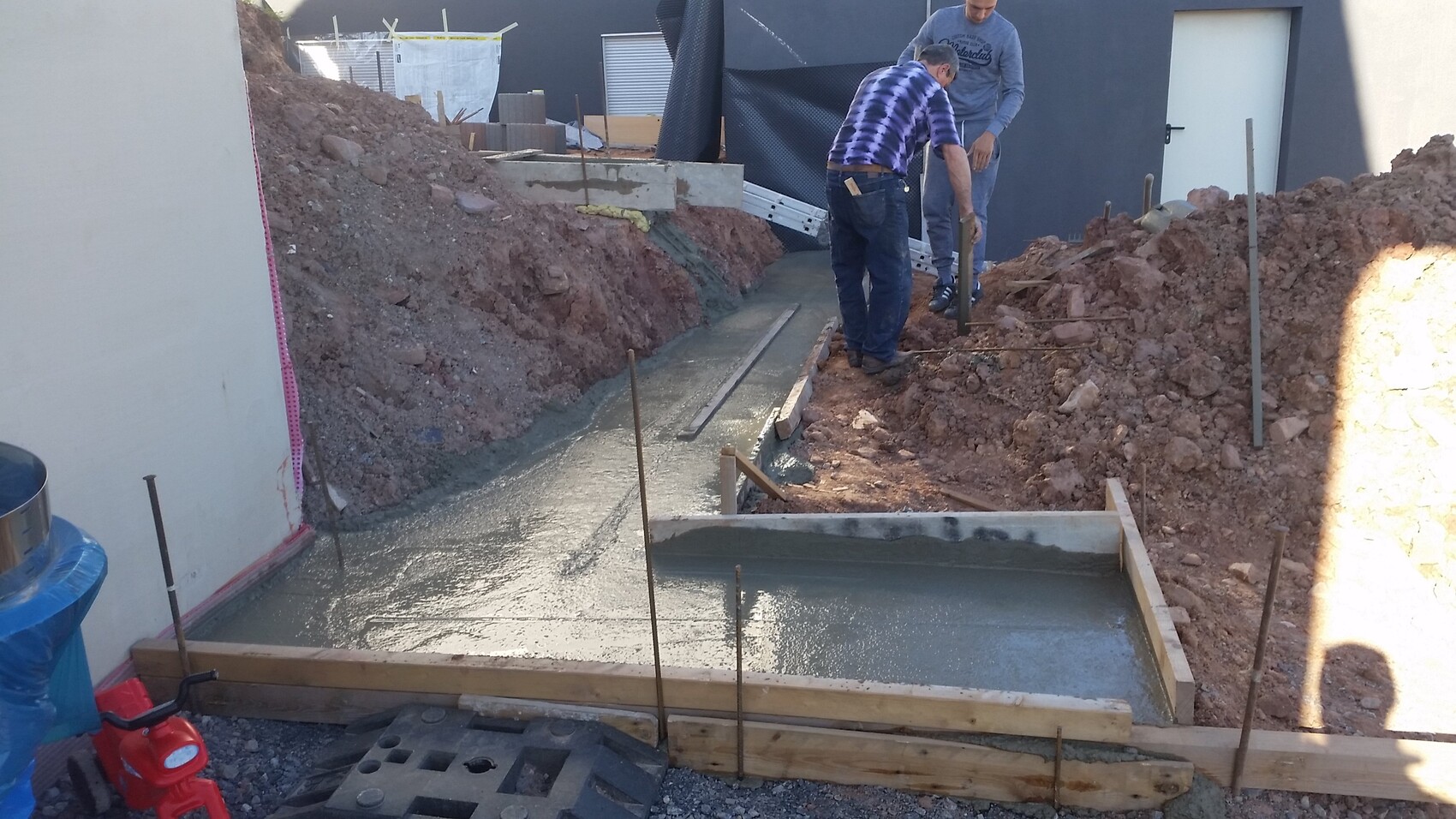 Two construction workers pouring fresh concrete foundation into a wooden trench form, with reinforcement bars.