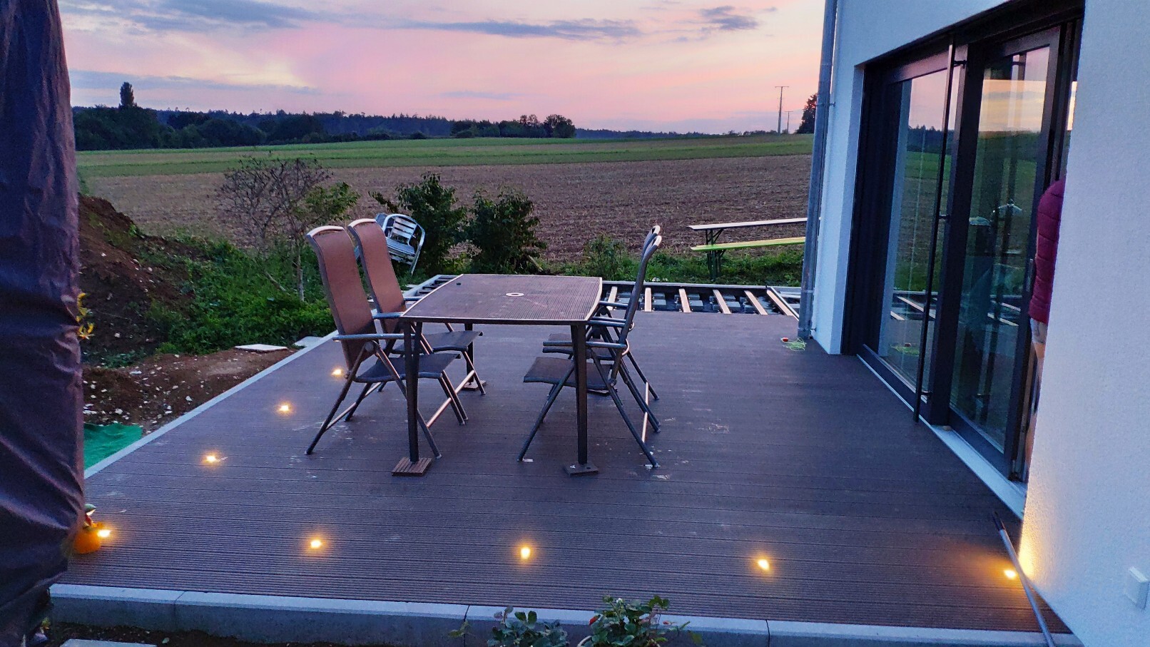 Evening wooden terrace with dining table and chairs, view of fields behind the house.