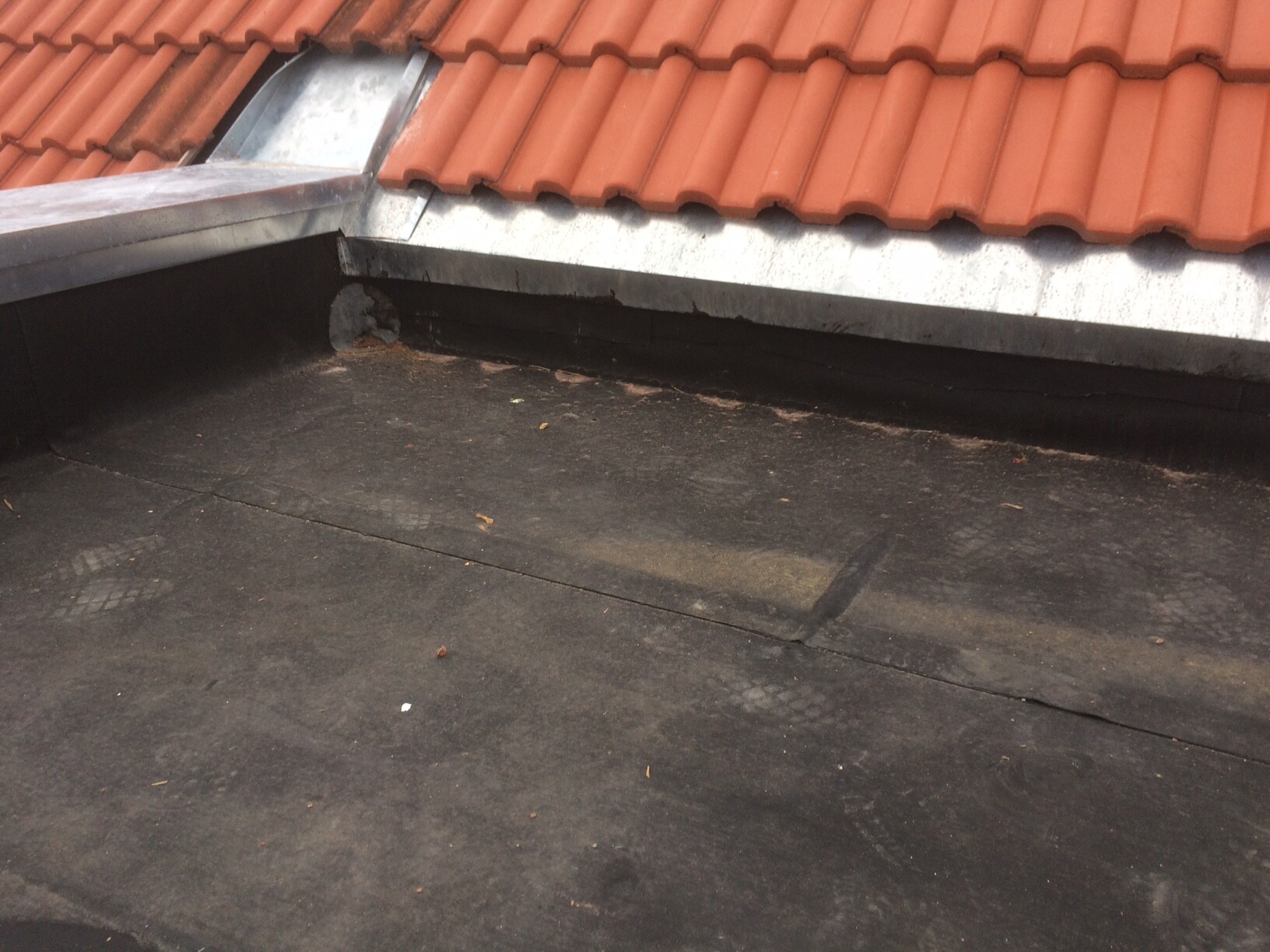 Roof surface with red clay tiles at the top edge and black bitumen membrane below; dust.