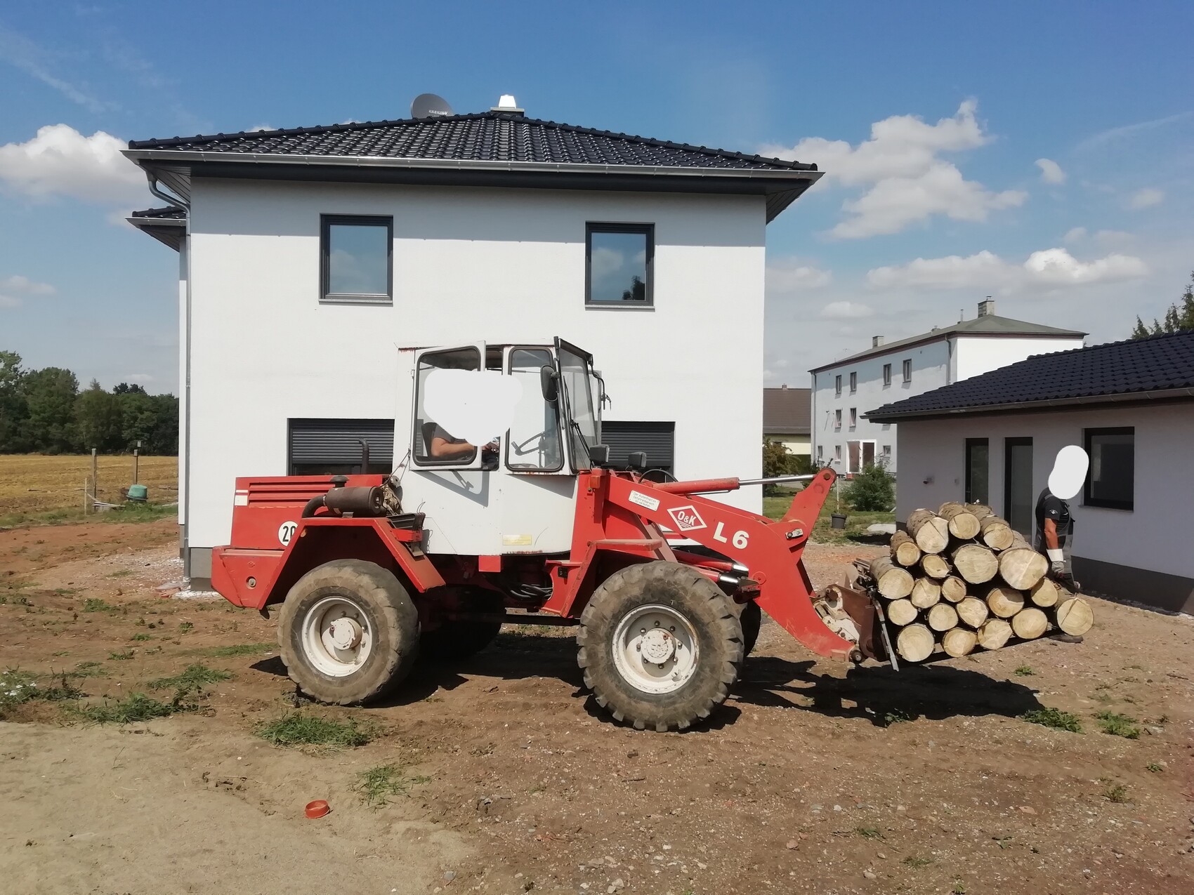Red front loader lifting a wood stack in front of a white two-story house; man standing beside it.