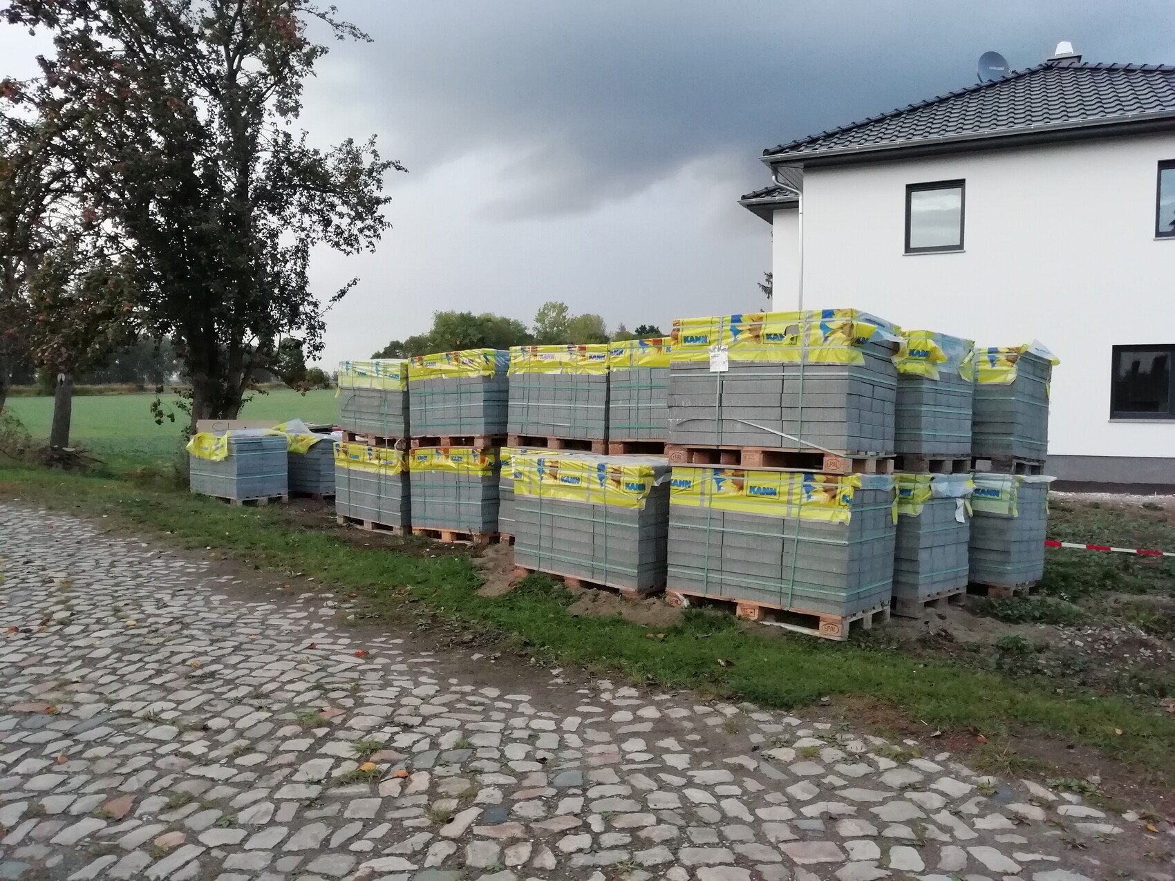 Several pallets of concrete roof tiles at the edge of a white house on cobblestone path.