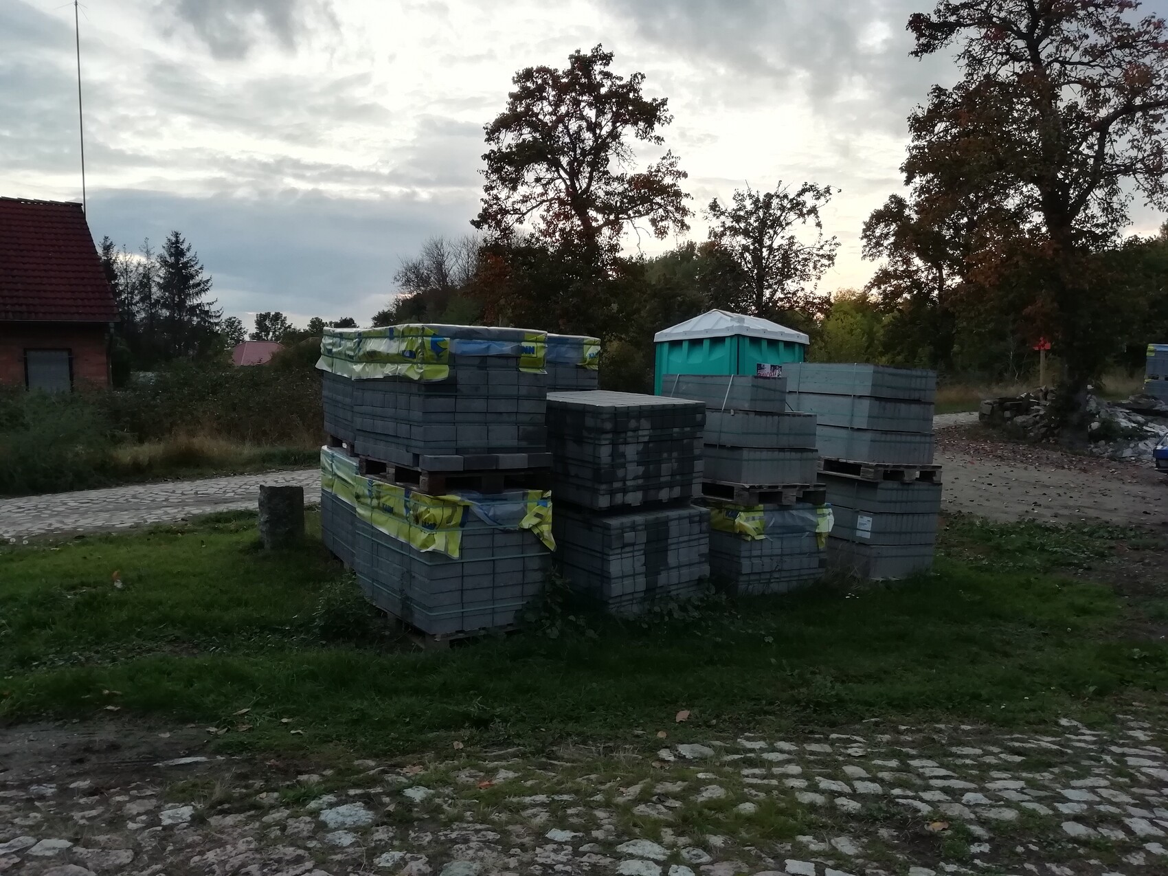 Stack of concrete slabs on pallets next to a portable toilet, rows of trees in the background.