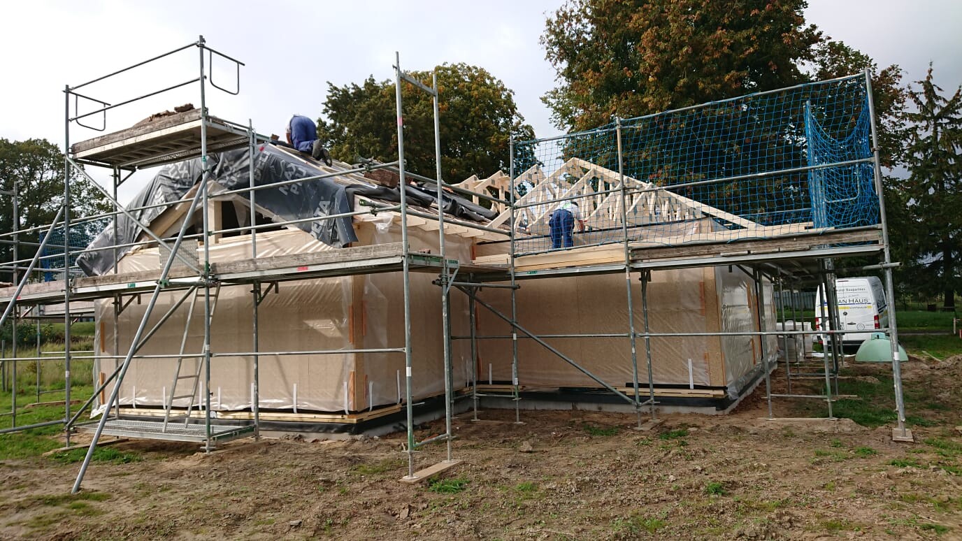 Construction site with scaffolding: wooden house structure, roof framework visible, protective net at the edge.