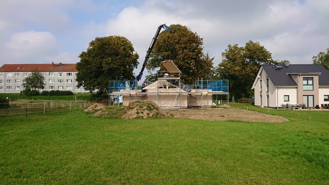 Crane lifting a wooden component onto a construction with scaffolding; green meadow and houses in the background.