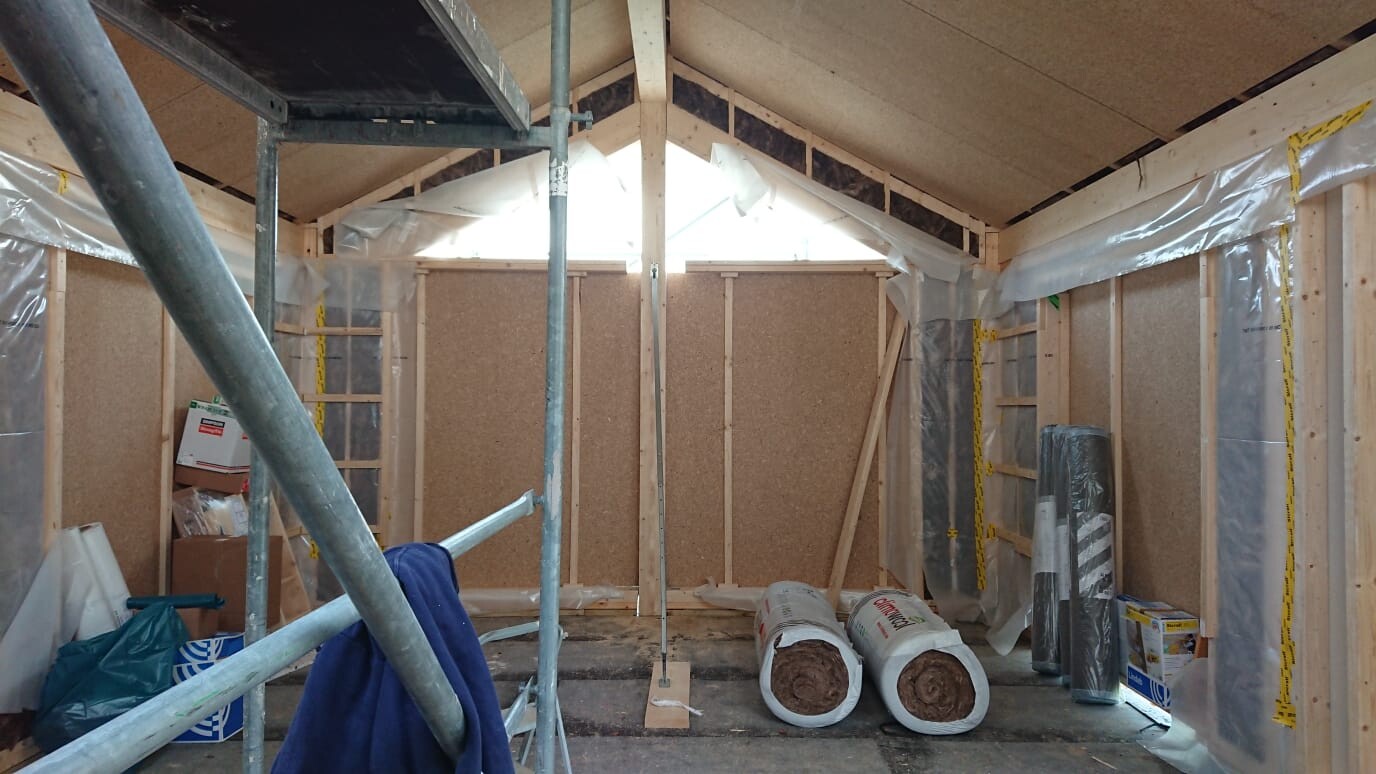 Construction setup: wooden walls, plastic sheeting, scaffolding poles, insulation rolls in an unfinished room.