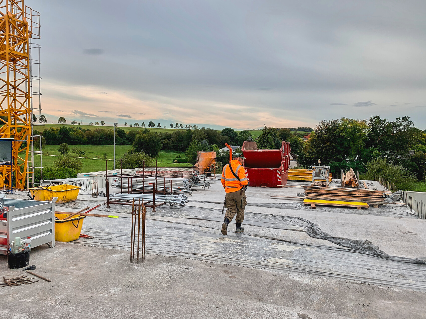 Construction site: worker in safety vest in front of concrete slab; reinforcement, crane on the left, green landscape.