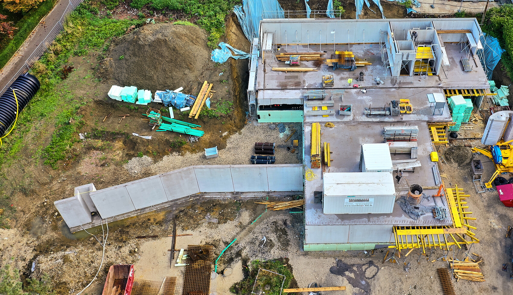 Bird’s eye view of a construction site with unfinished building, material stacks, railing, and construction machinery.