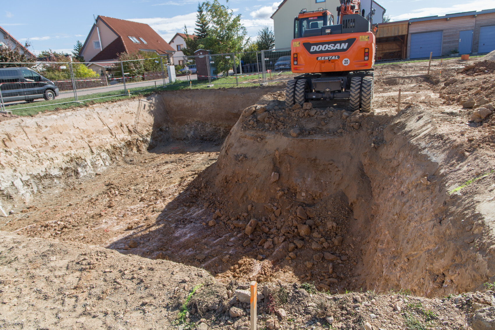 Große Baugrube auf einer Baustelle, ein orangefarbener Doosan-Bagger am Rand, Häuser im Hintergrund.