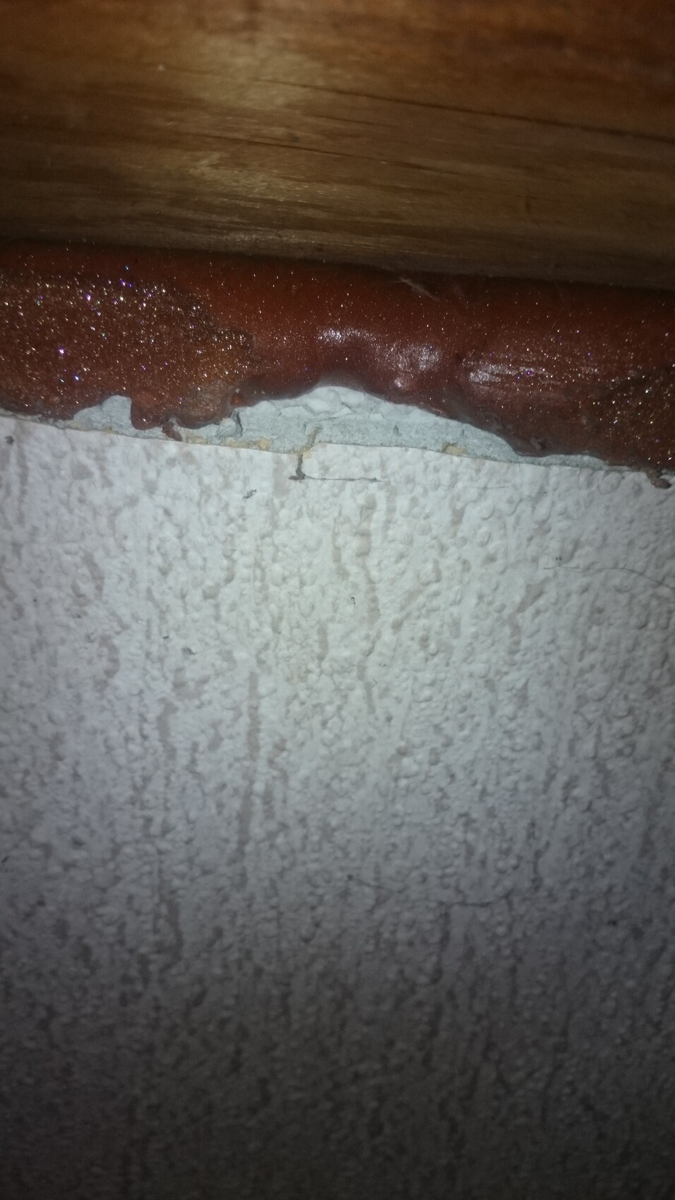 Reddish-brown sticky substance on the edge between a wooden strip and a rough white wall.