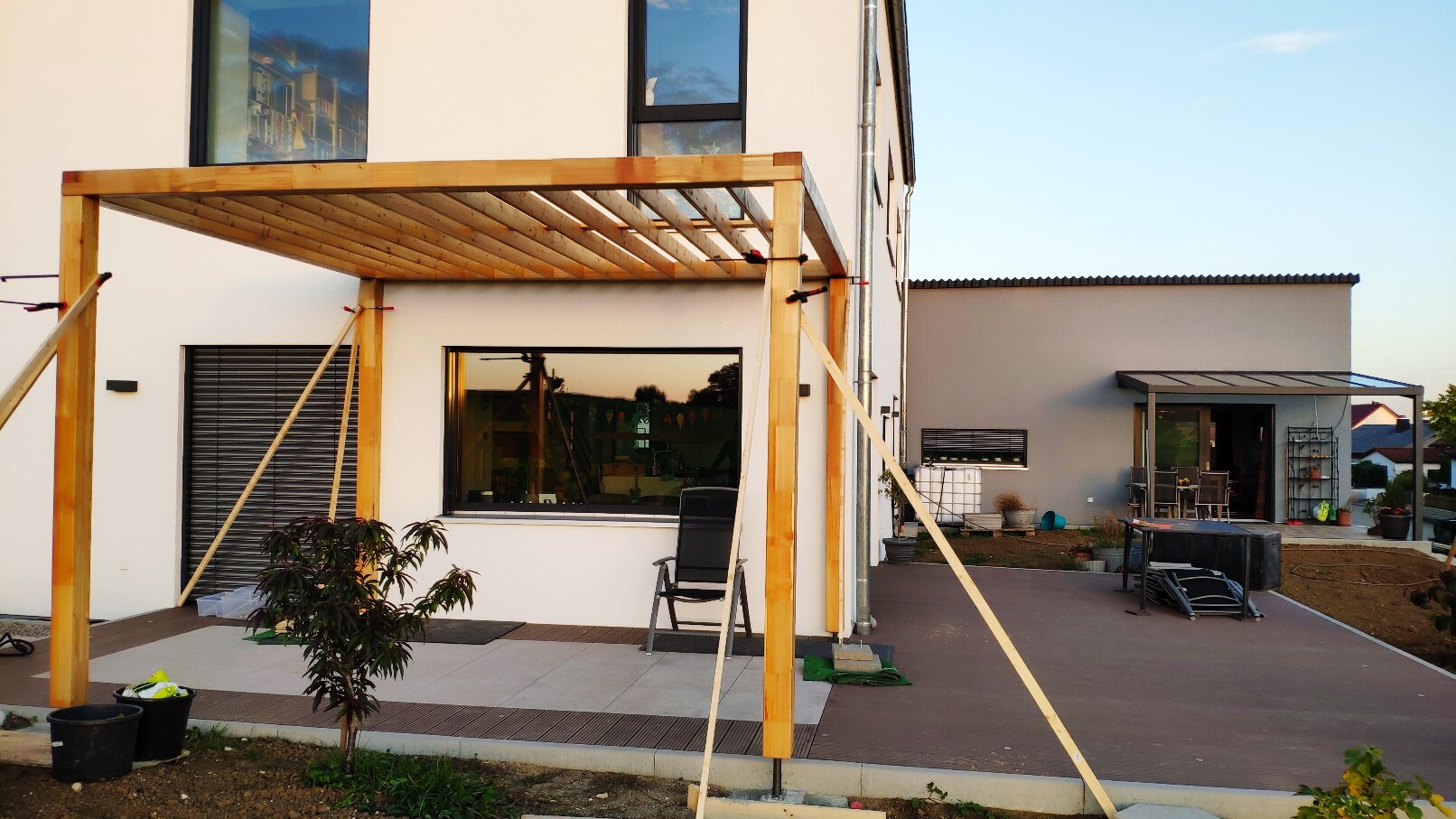 Holz-Pergola wird am Haus aufgebaut; Terrasse mit Stuhl, Pflanze und Gartenbereich.