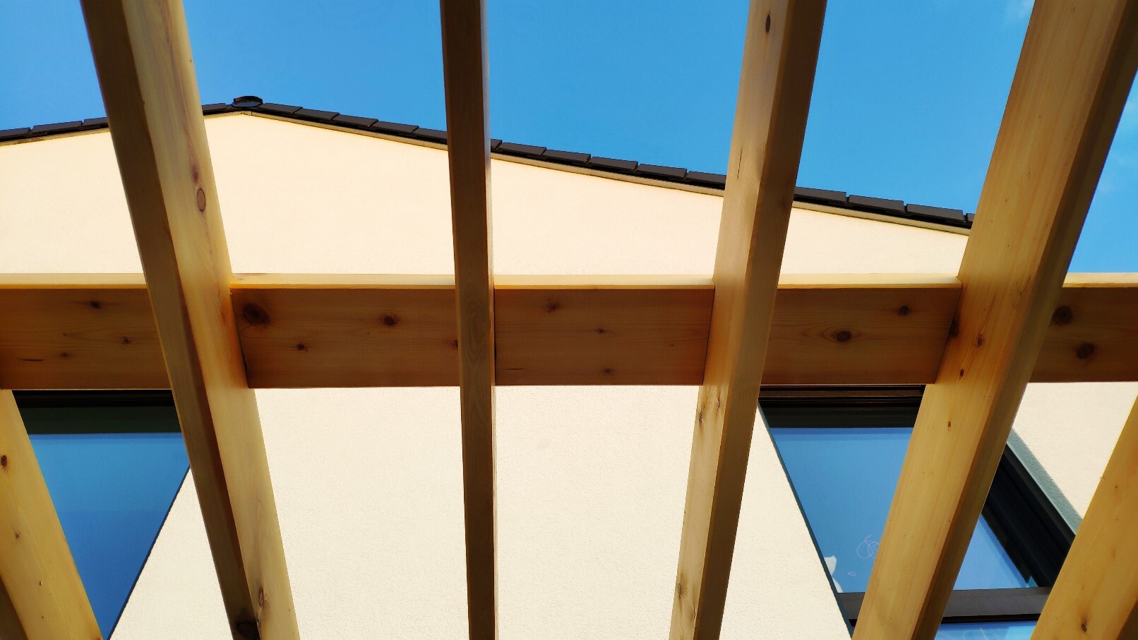Blick von unten durch eine Holzkonstruktion auf eine helle Fassade mit Fenstern und blauem Himmel.