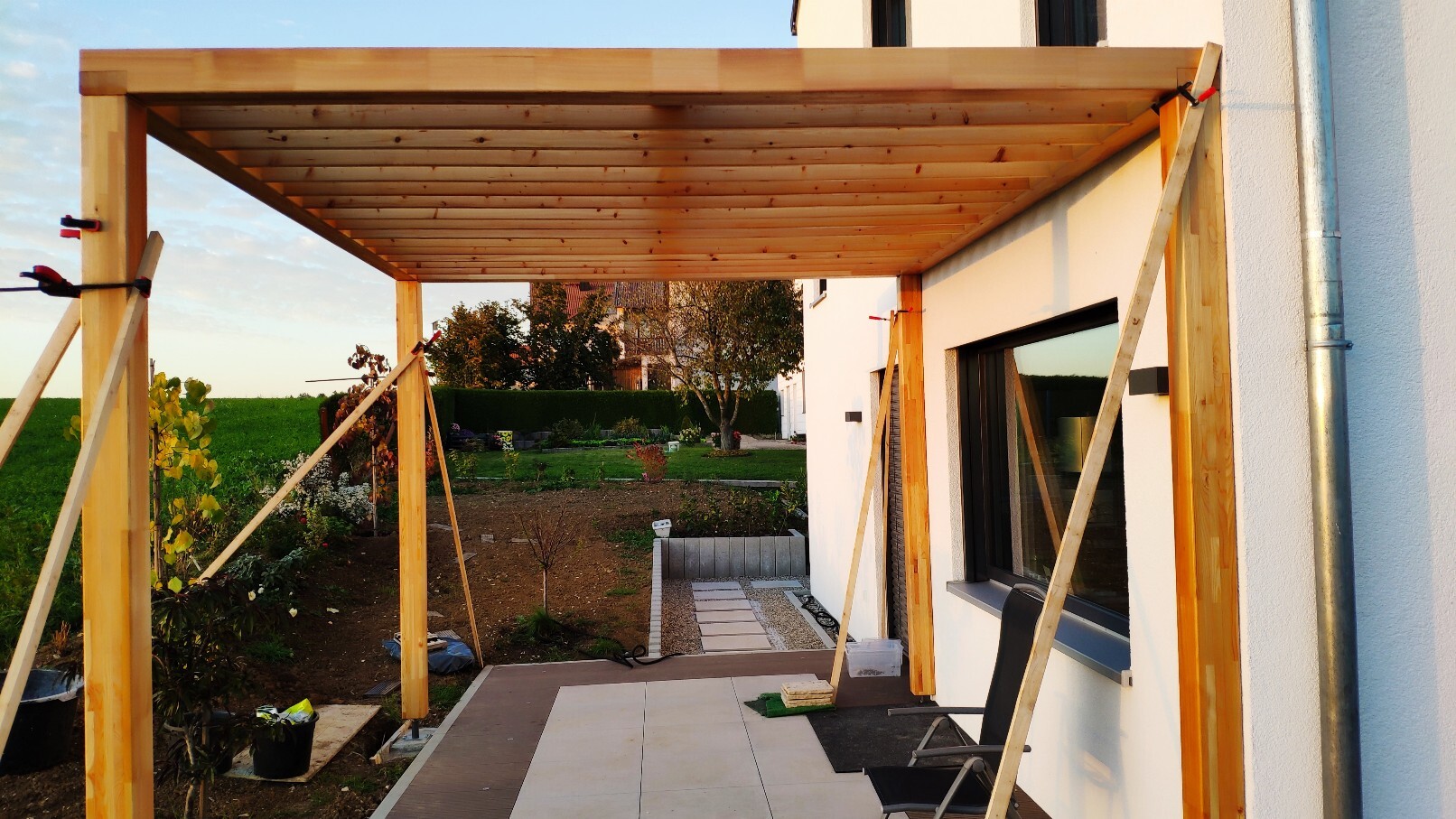 Holz-Pergola vor weißem Haus mit Terrasse; Garten im Hintergrund.